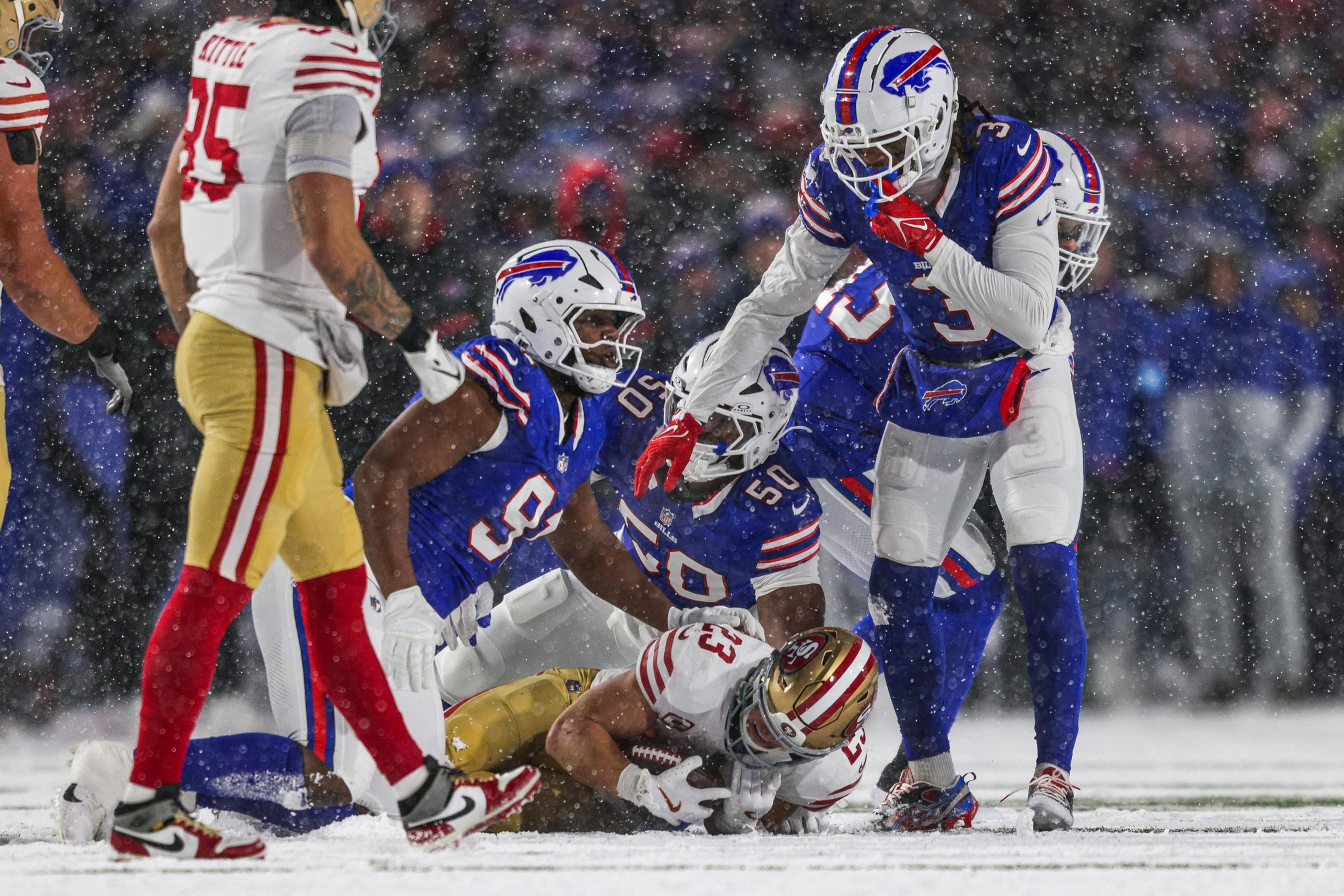 Football players in blue uniforms tackle a player in a white uniform holding the ball, on a snow-covered field. Snow is visibly falling around them.