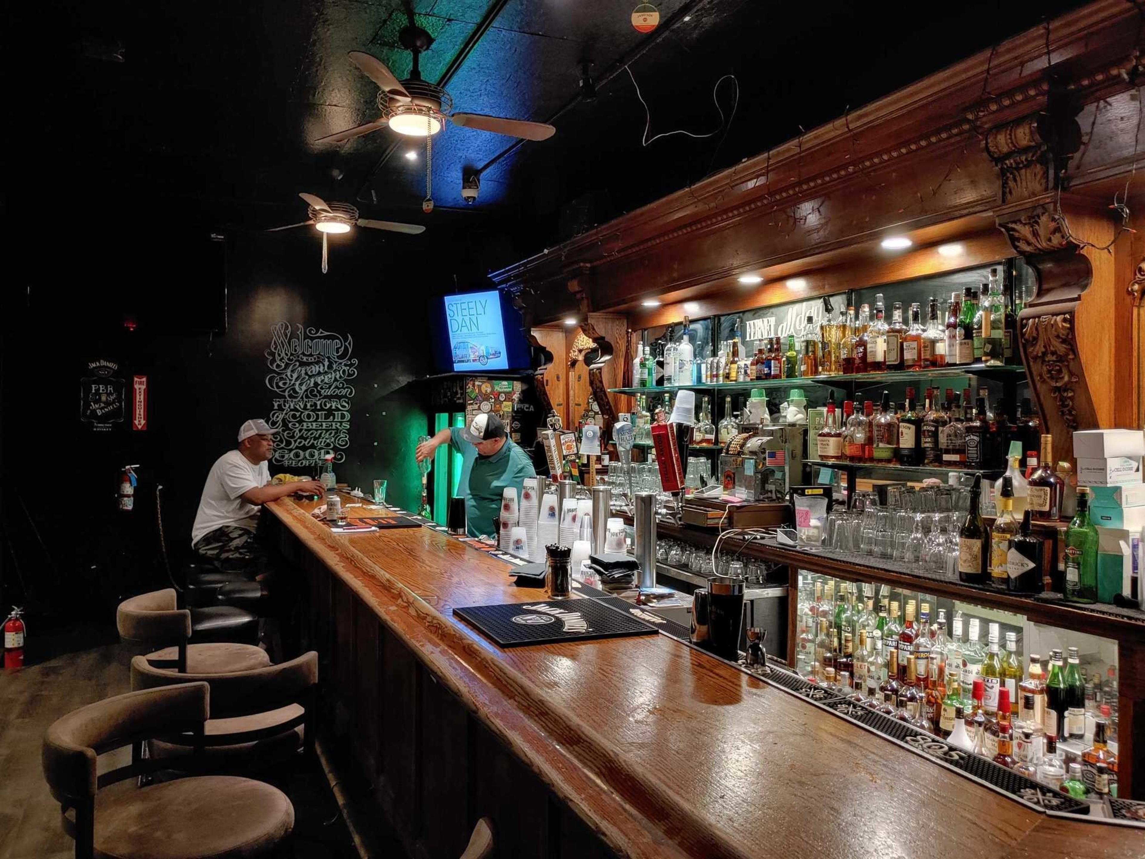 A long wooden bar with bottles and glasses behind it, two men wearing caps and casual clothes are engaged in conversation.