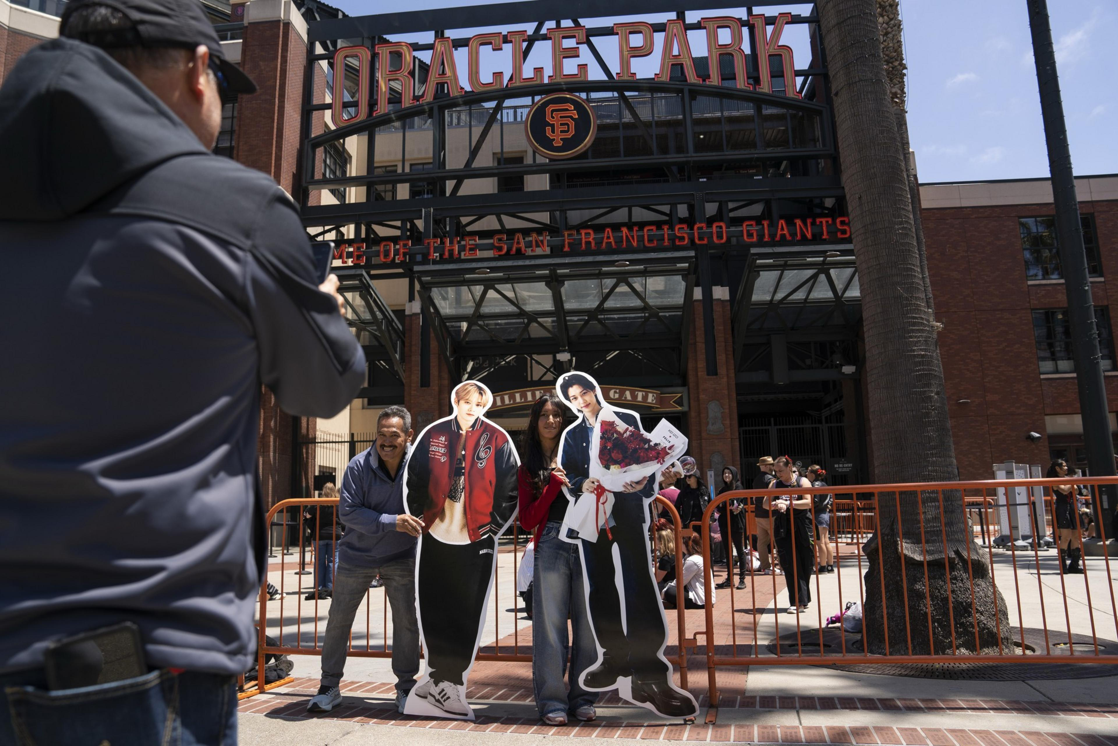 Two people pose with cutouts of stars outside Oracle Park, San Francisco. A third person is taking their photo. An orange barricade surrounds them.