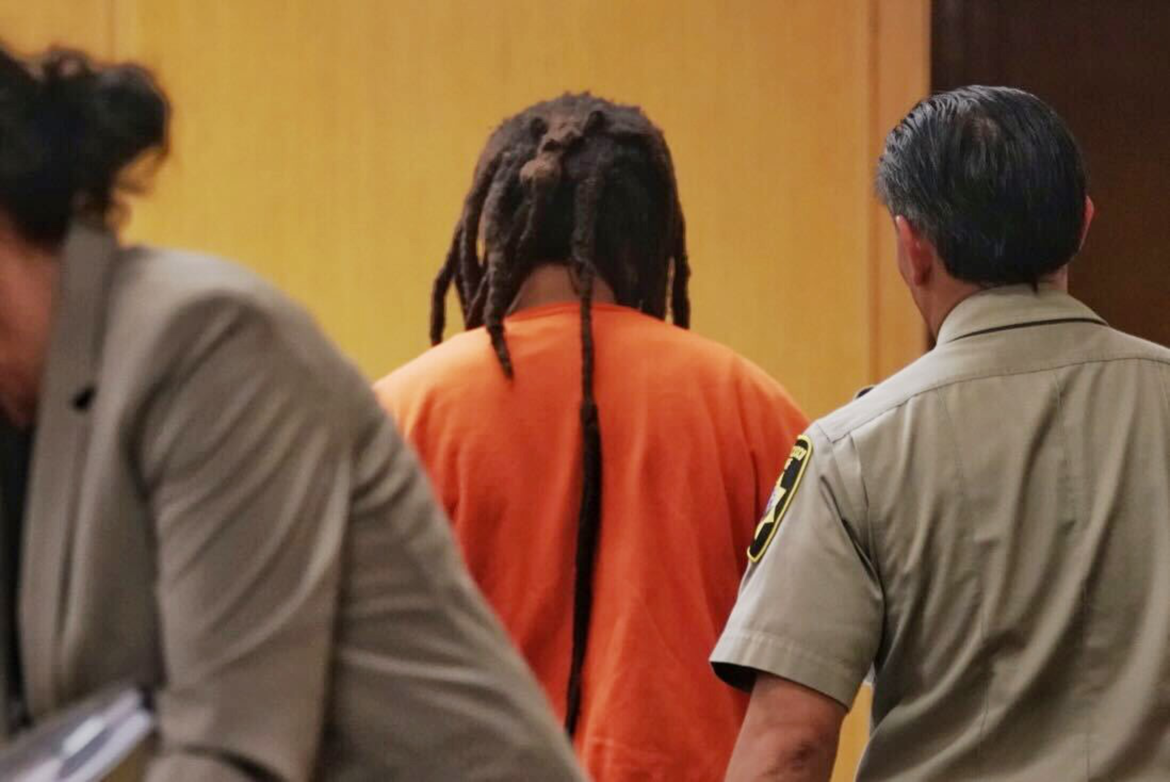A view of a back of man in an orange jumpsuit walking away from the camera being led by a man in a sheriff's uniform.