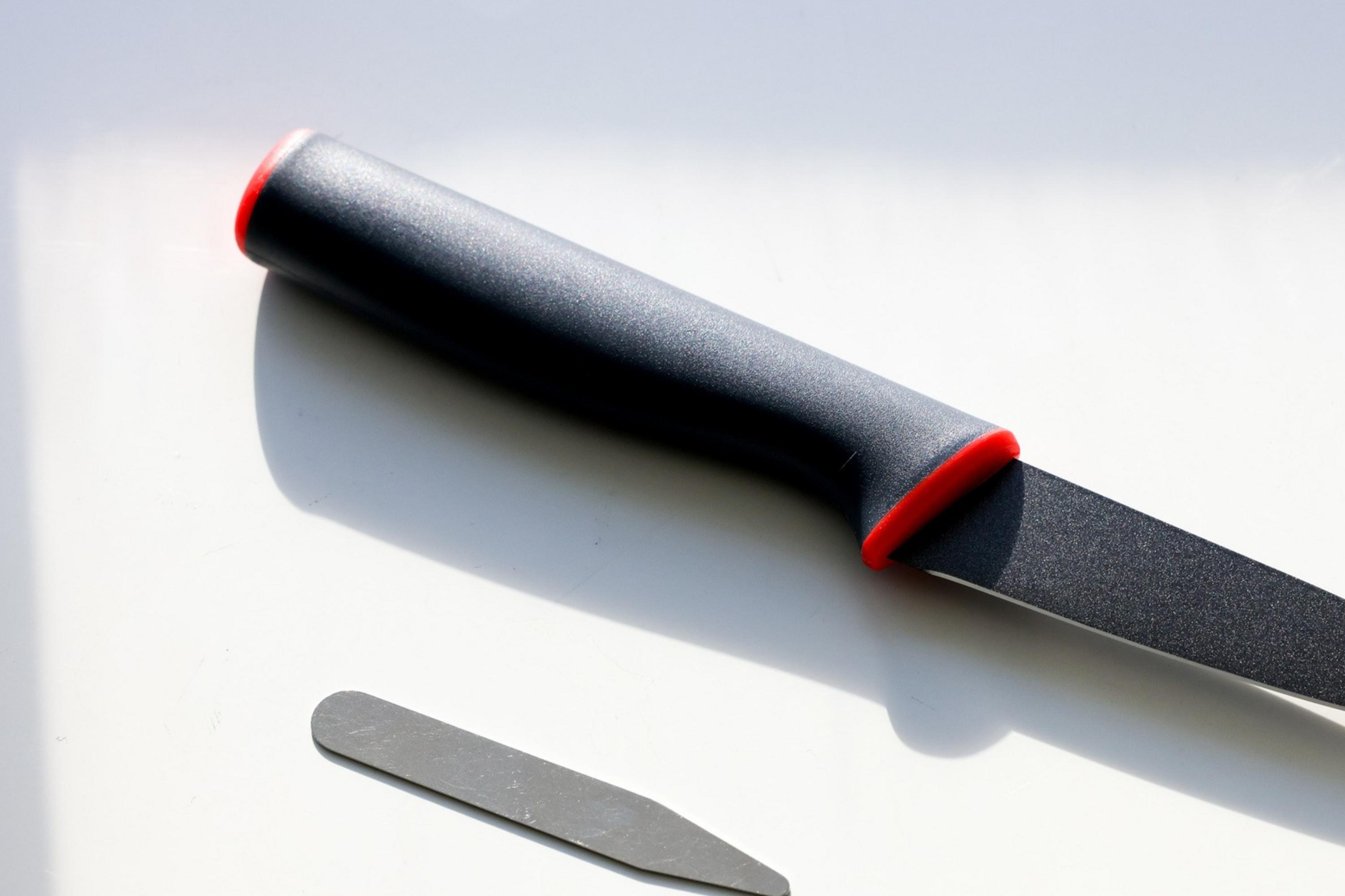 A black knife with a red accent on the handle is placed on a light surface, casting a shadow. Nearby, a small, flat metal collar stay is also in the image.