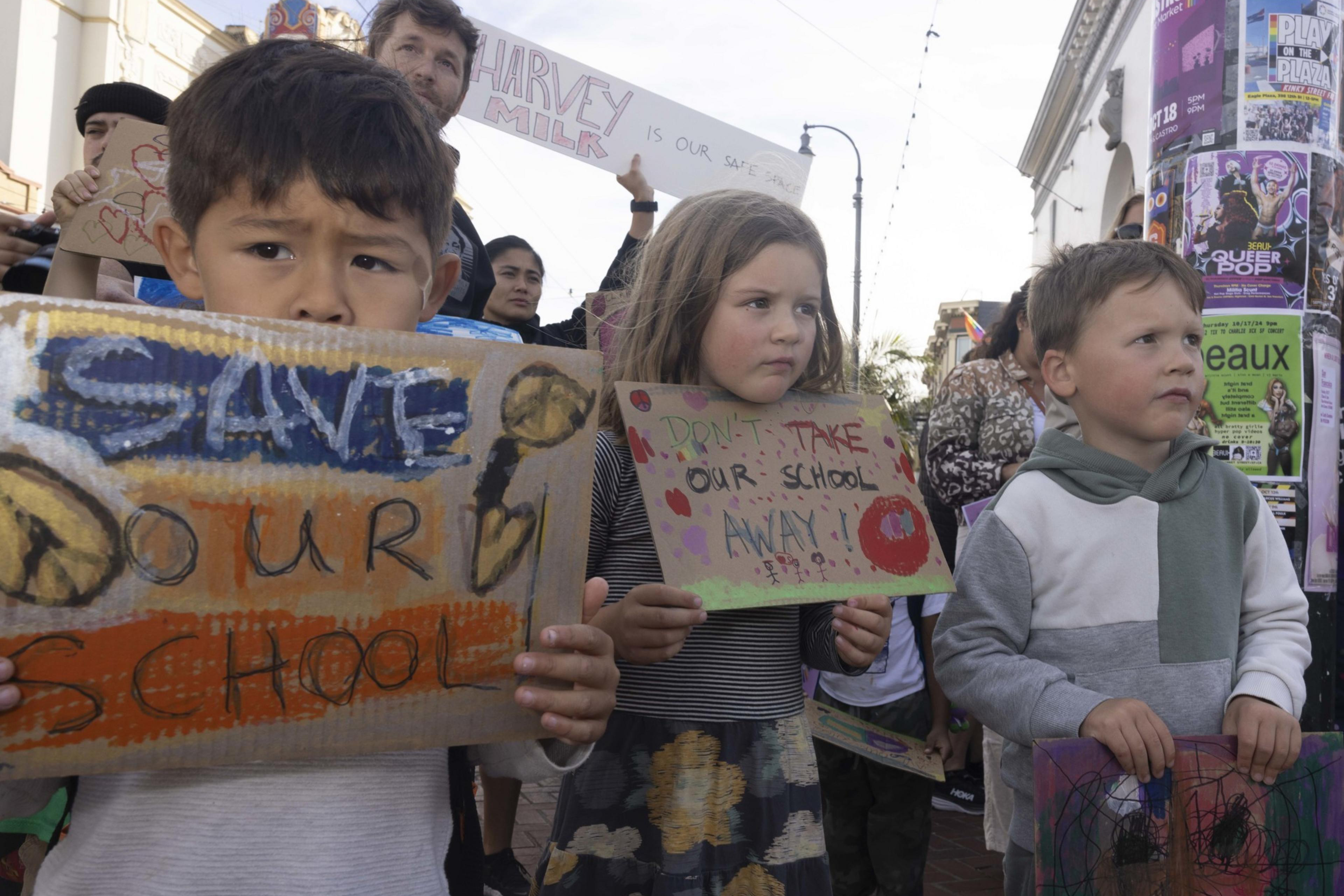 Children hold handmade signs during a protest. Their signs say â€œSave Our Schoolâ€ and â€œDonâ€™t Take Our School Away!â€ amidst a backdrop of urban posters.