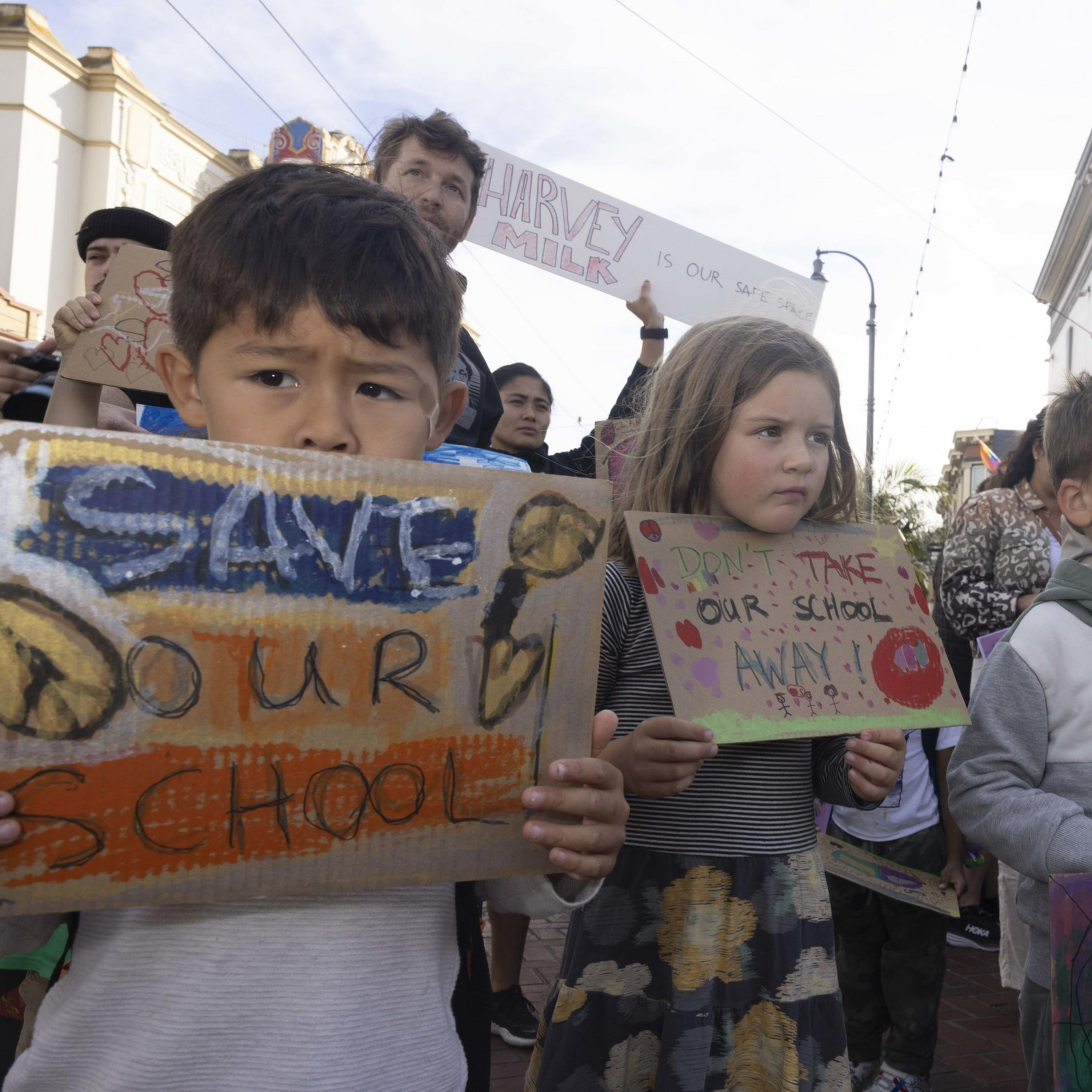 Children hold handmade signs during a protest. Their signs say â€œSave Our Schoolâ€ and â€œDonâ€™t Take Our School Away!â€ amidst a backdrop of urban posters.