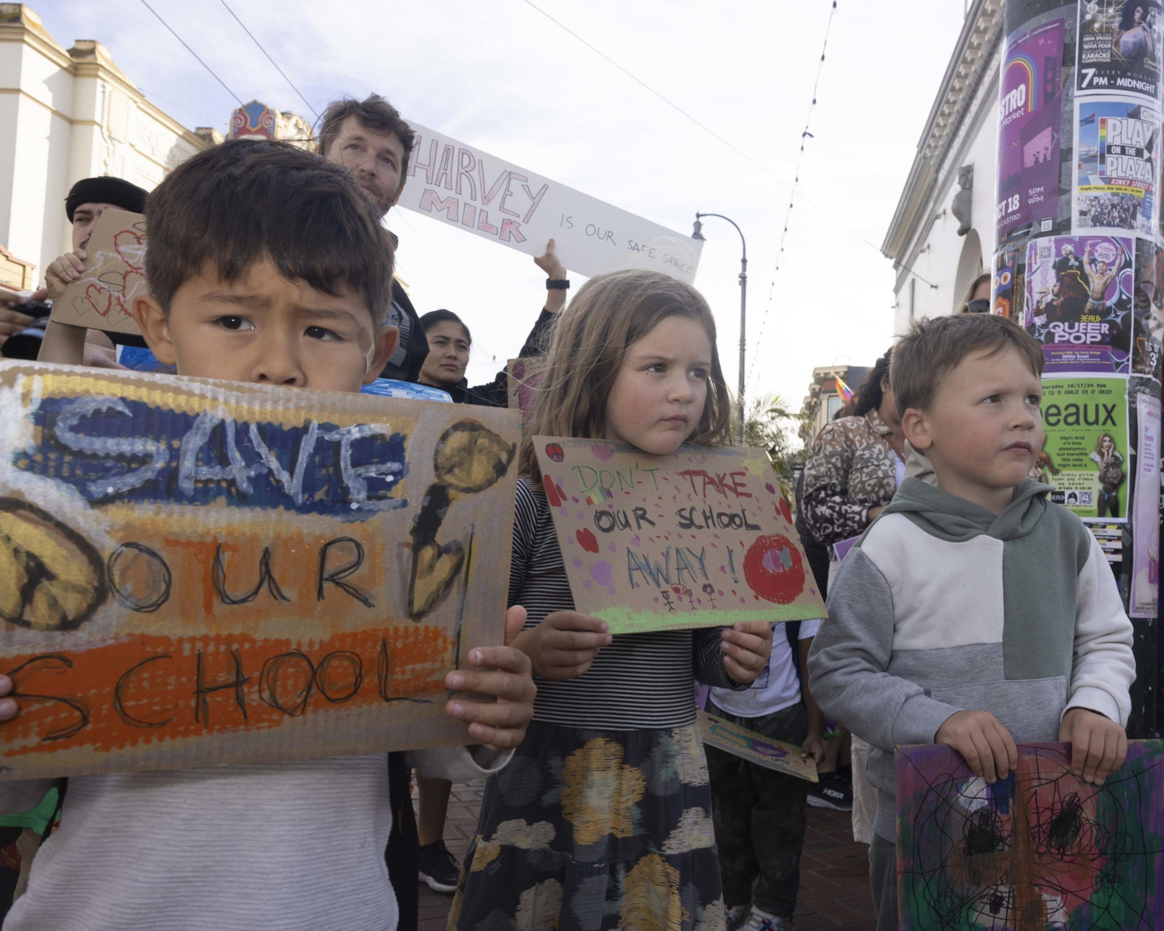 Children hold handmade signs during a protest. Their signs say â€œSave Our Schoolâ€ and â€œDonâ€™t Take Our School Away!â€ amidst a backdrop of urban posters.