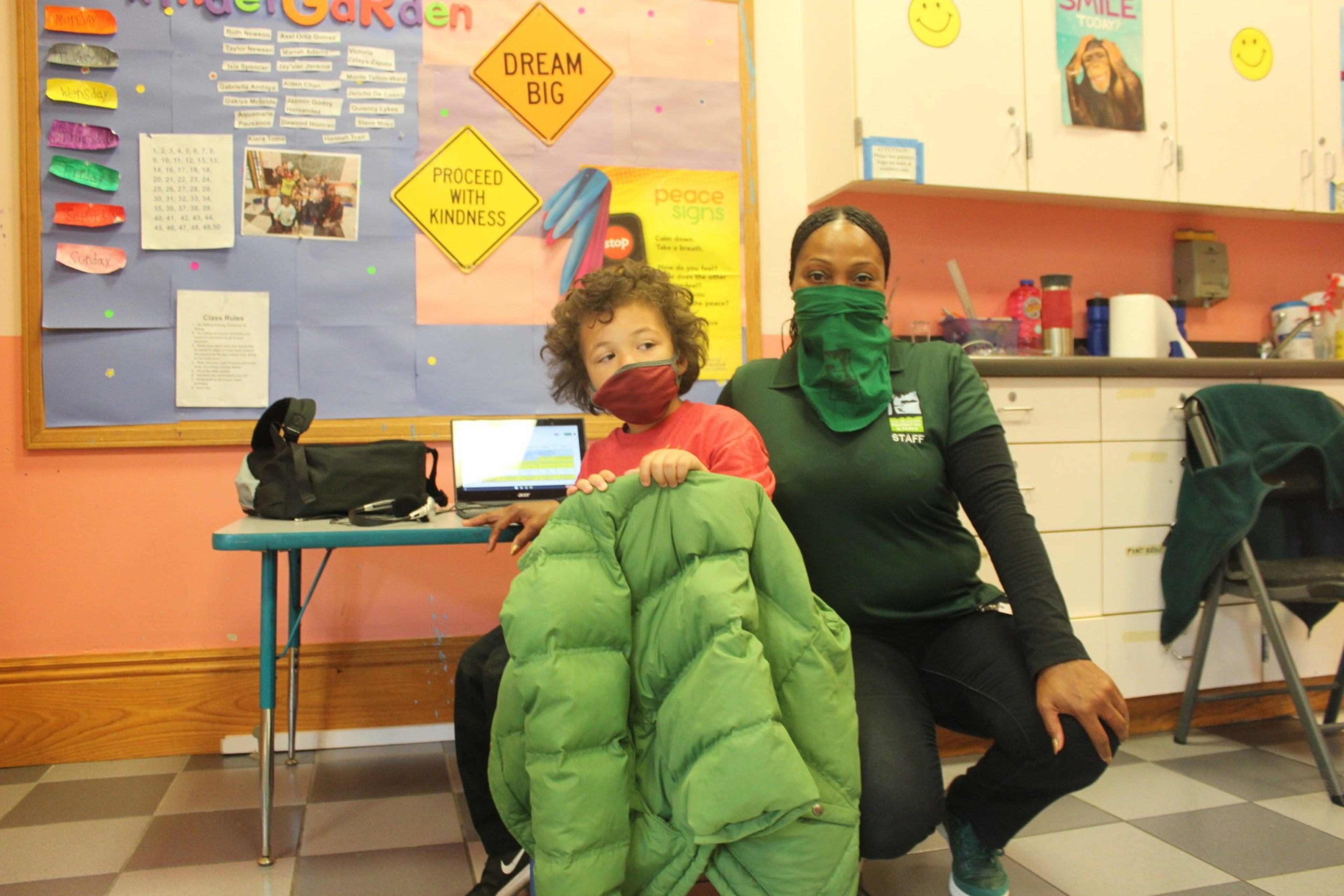 A young child wearing a red mask holds a green jacket, sitting next to a staff member in a green shirt and mask in a colorful classroom.