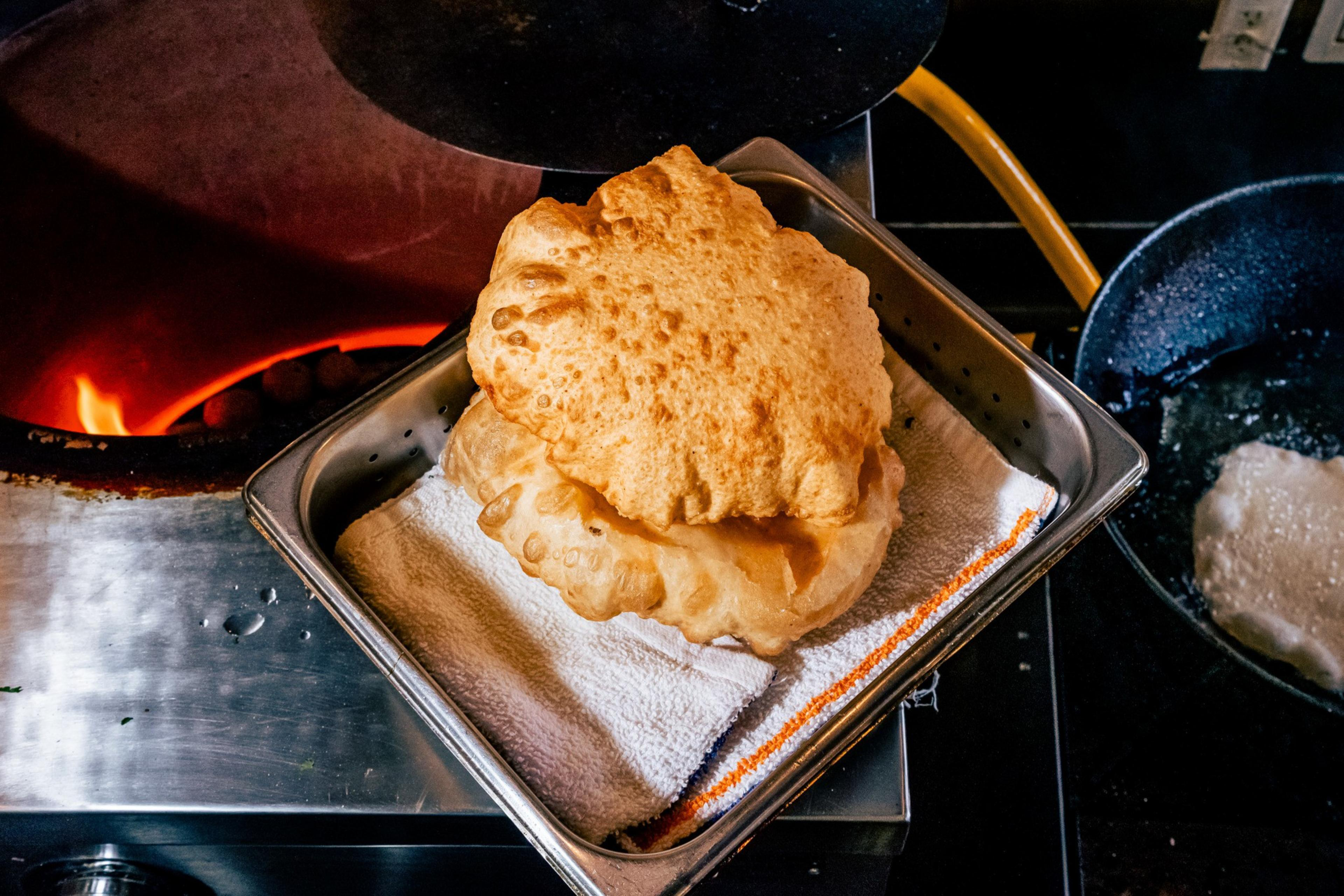 Two pieces of golden-brown, puffed-up fried bread resting on a towel-lined metal tray near a stove and frying pan.