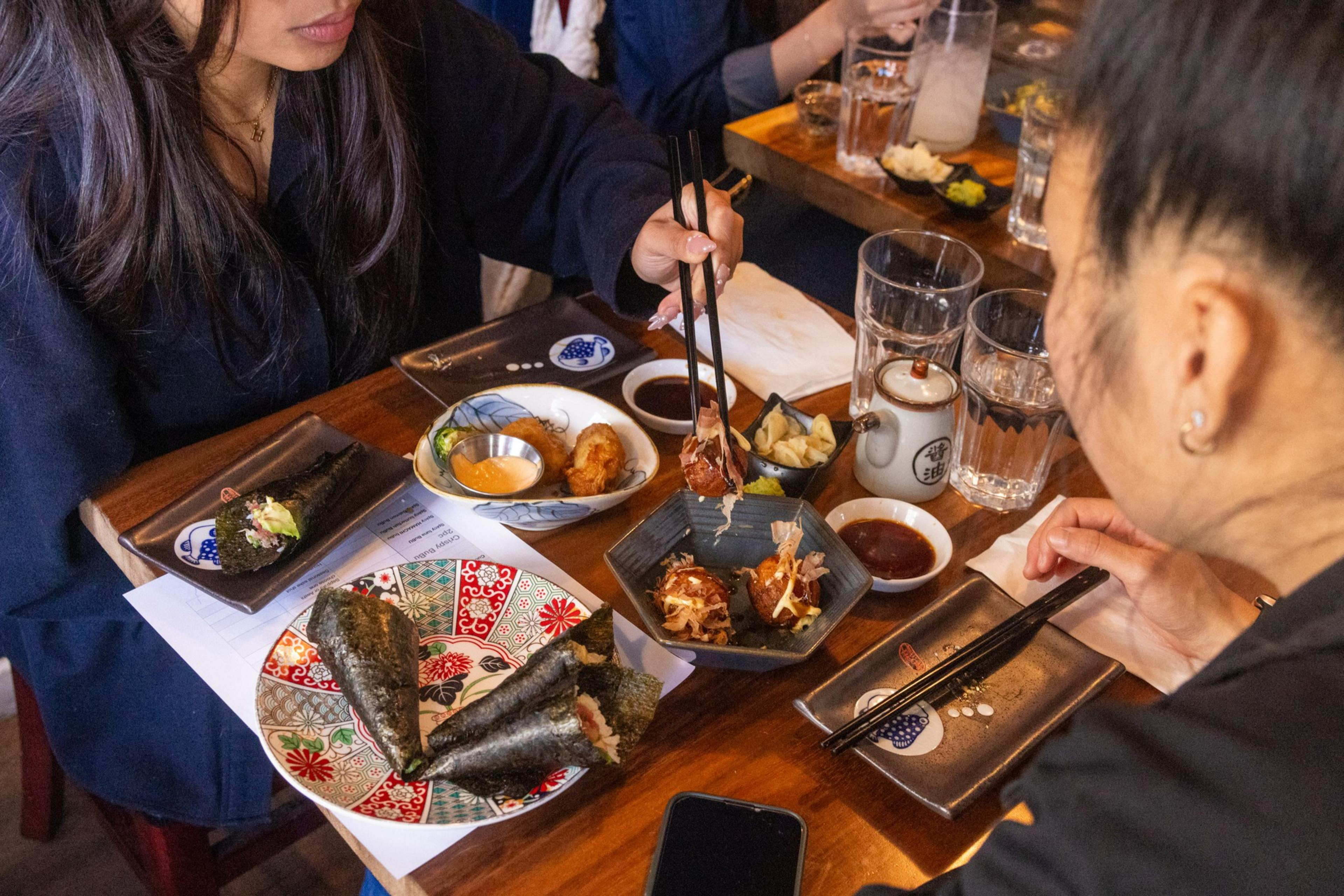 Two people are dining at a table with Japanese food, including hand rolls and takoyaki. Chopsticks and sauce dishes are present along with tea.