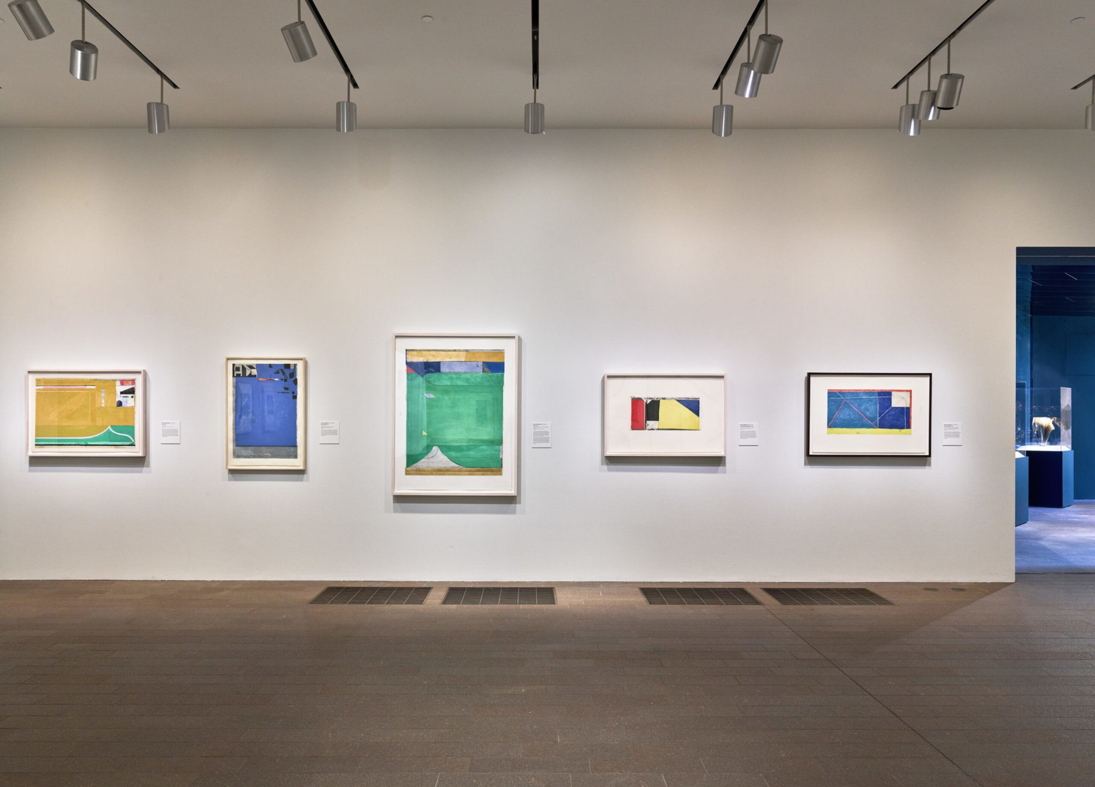 Five framed abstract paintings with geometric shapes and bold colors like green, blue, yellow, and red hang on a white gallery wall under track lighting.