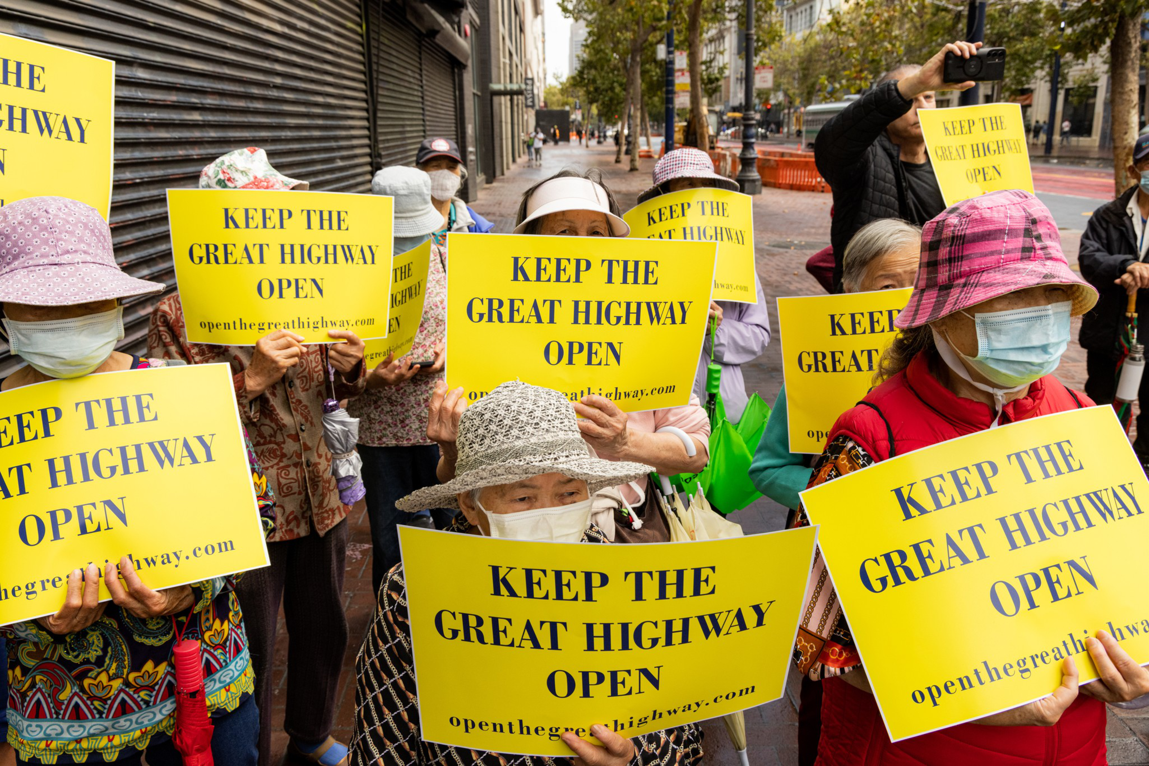 A group of people wearing hats and masks hold yellow signs reading &quot;KEEP THE GREAT HIGHWAY OPEN.&quot; They're gathered on a city street, showing their support for a cause.