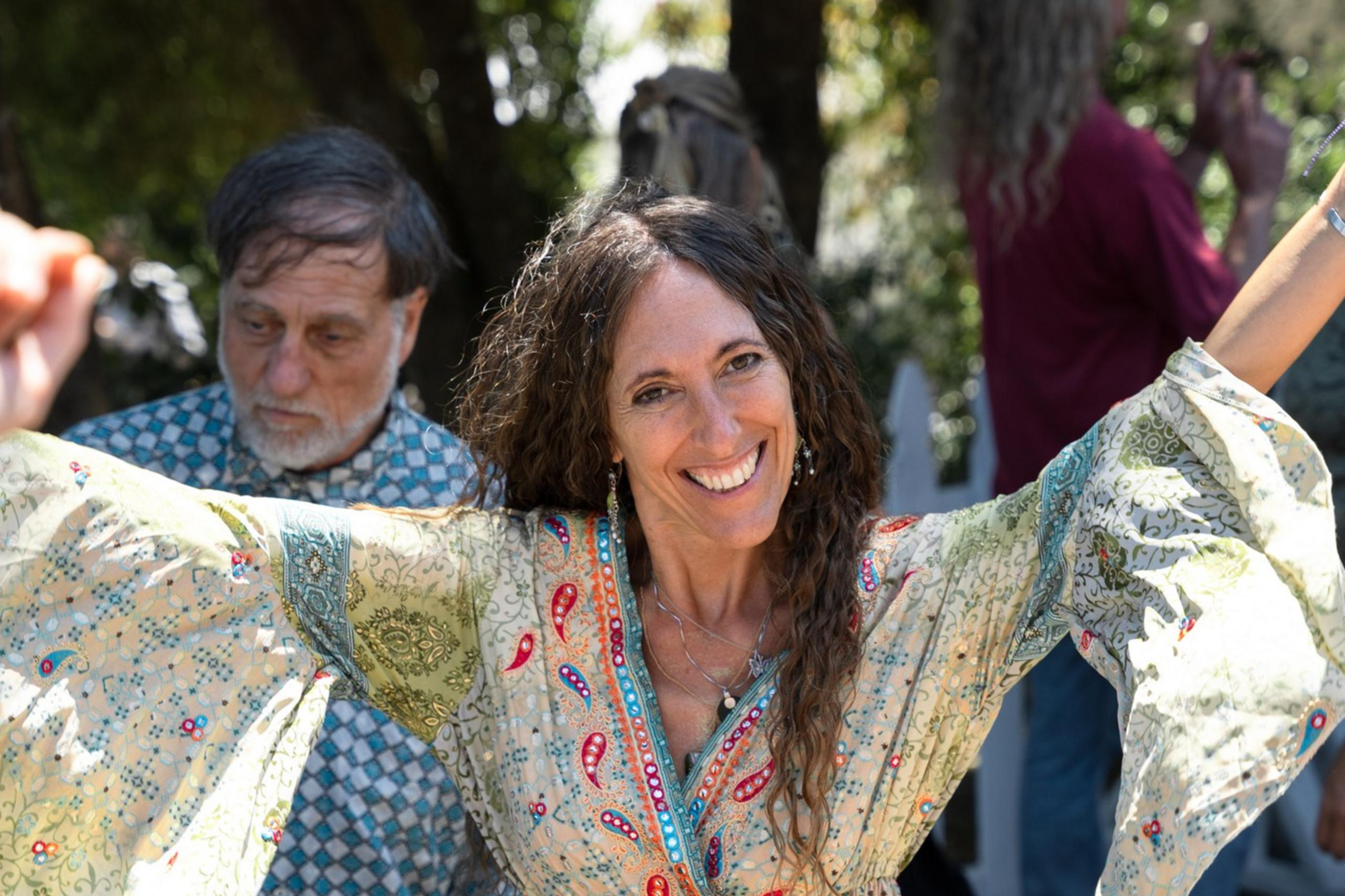 A woman with long curly hair smiles brightly and spreads her arms cheerfully in an outdoor setting. She wears a long, patterned dress with bell sleeves. Others are visible in the background.