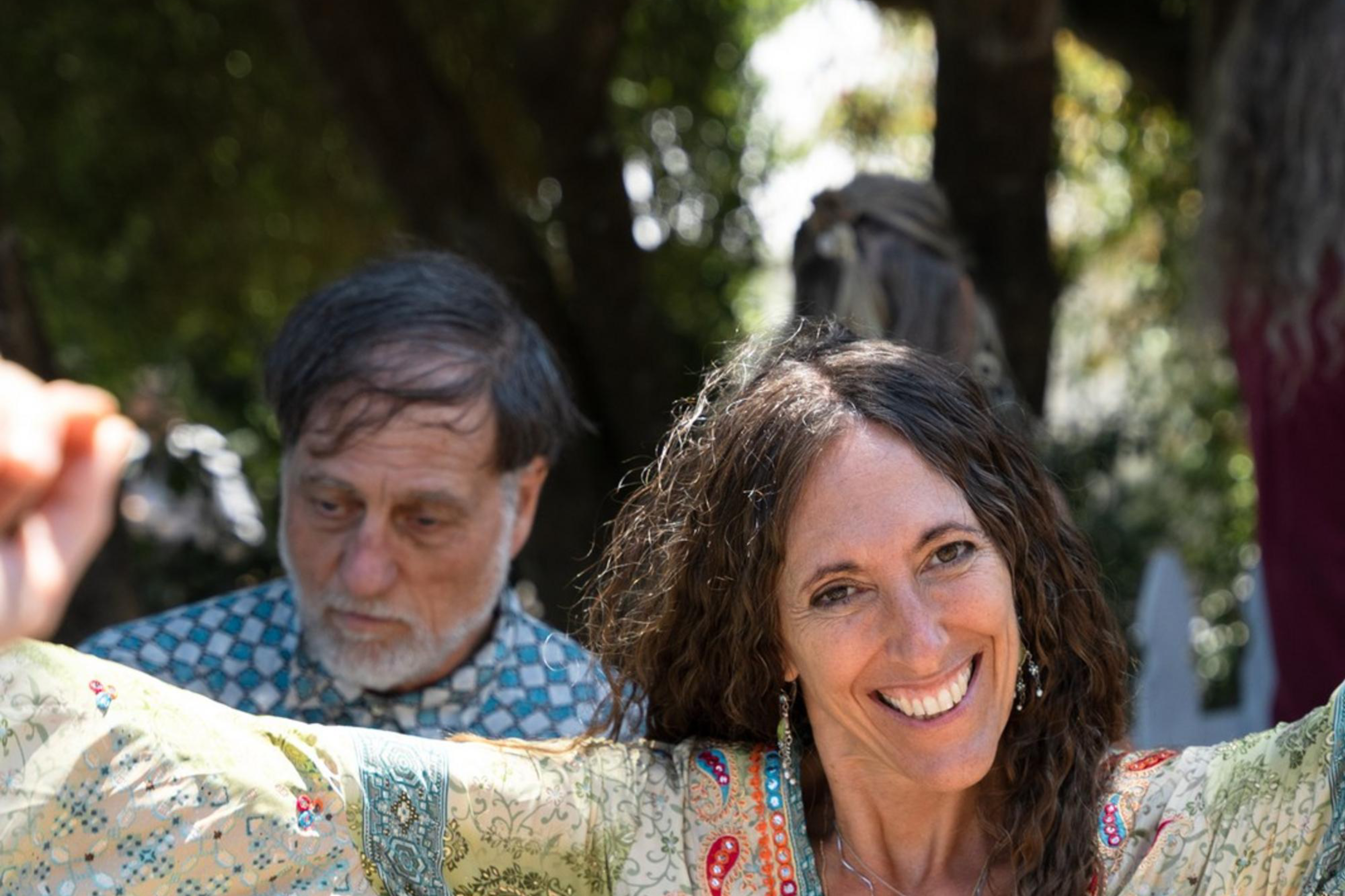 A woman with long curly hair smiles brightly and spreads her arms cheerfully in an outdoor setting. She wears a long, patterned dress with bell sleeves. Others are visible in the background.