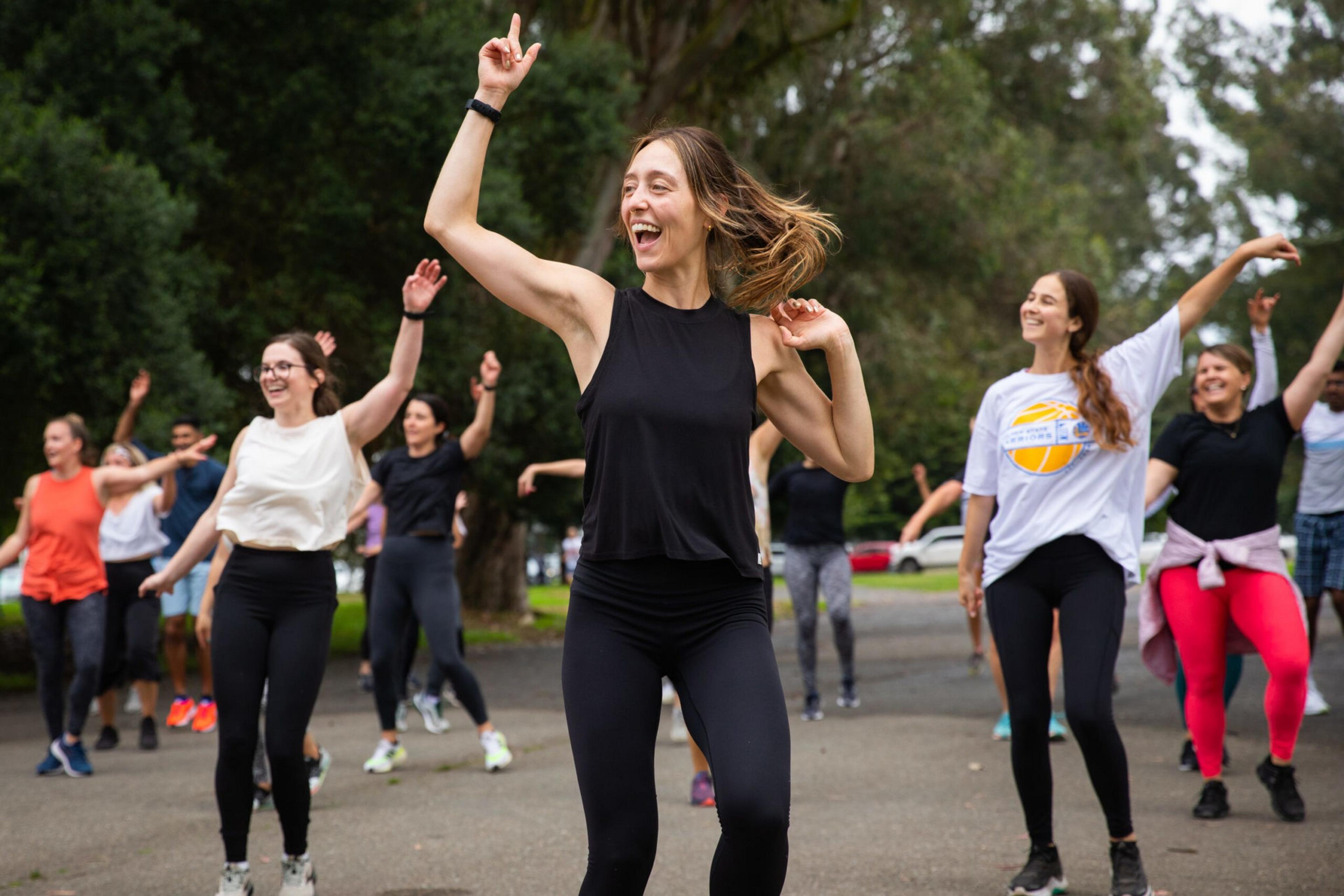 A group of people, mostly women, are dancing energetically outdoors on a paved path surrounded by trees, all smiling and enjoying themselves.