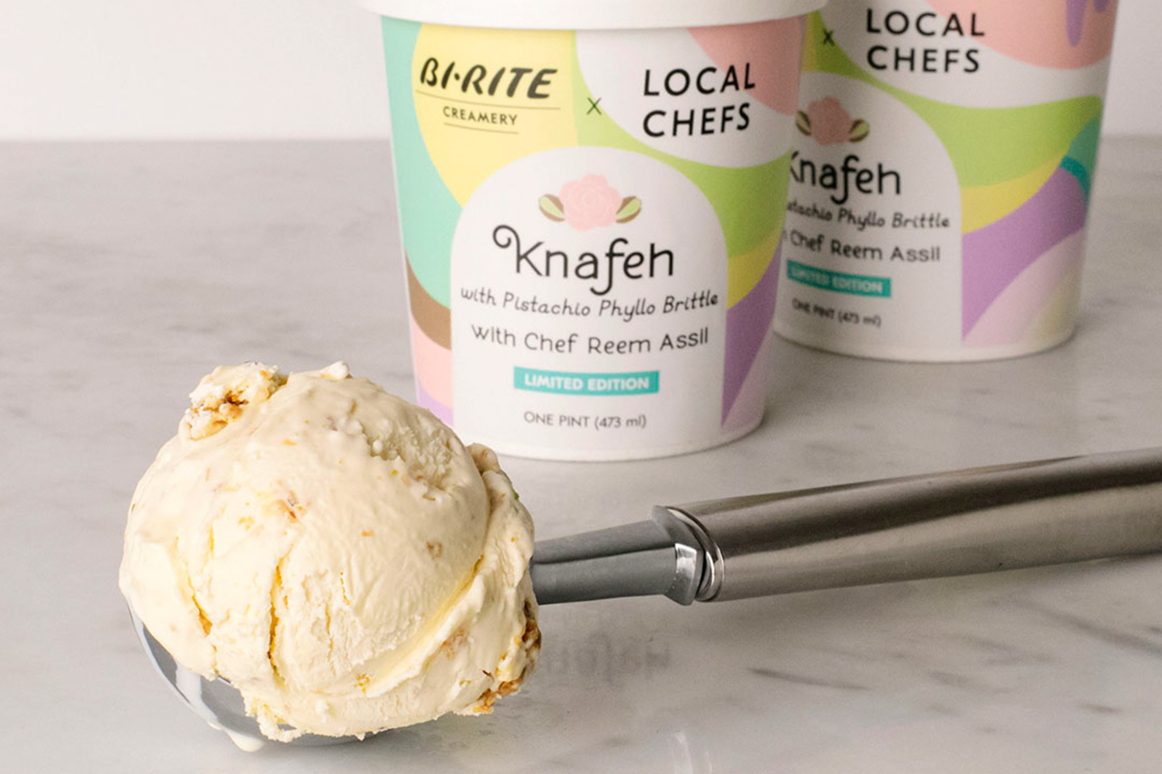 A scoop of Knafeh ice cream with pistachio phyllo brittle sits on a metal ice cream scoop, with two colorful ice cream tubs labeled "B-RITE Creamery x Local Chefs."