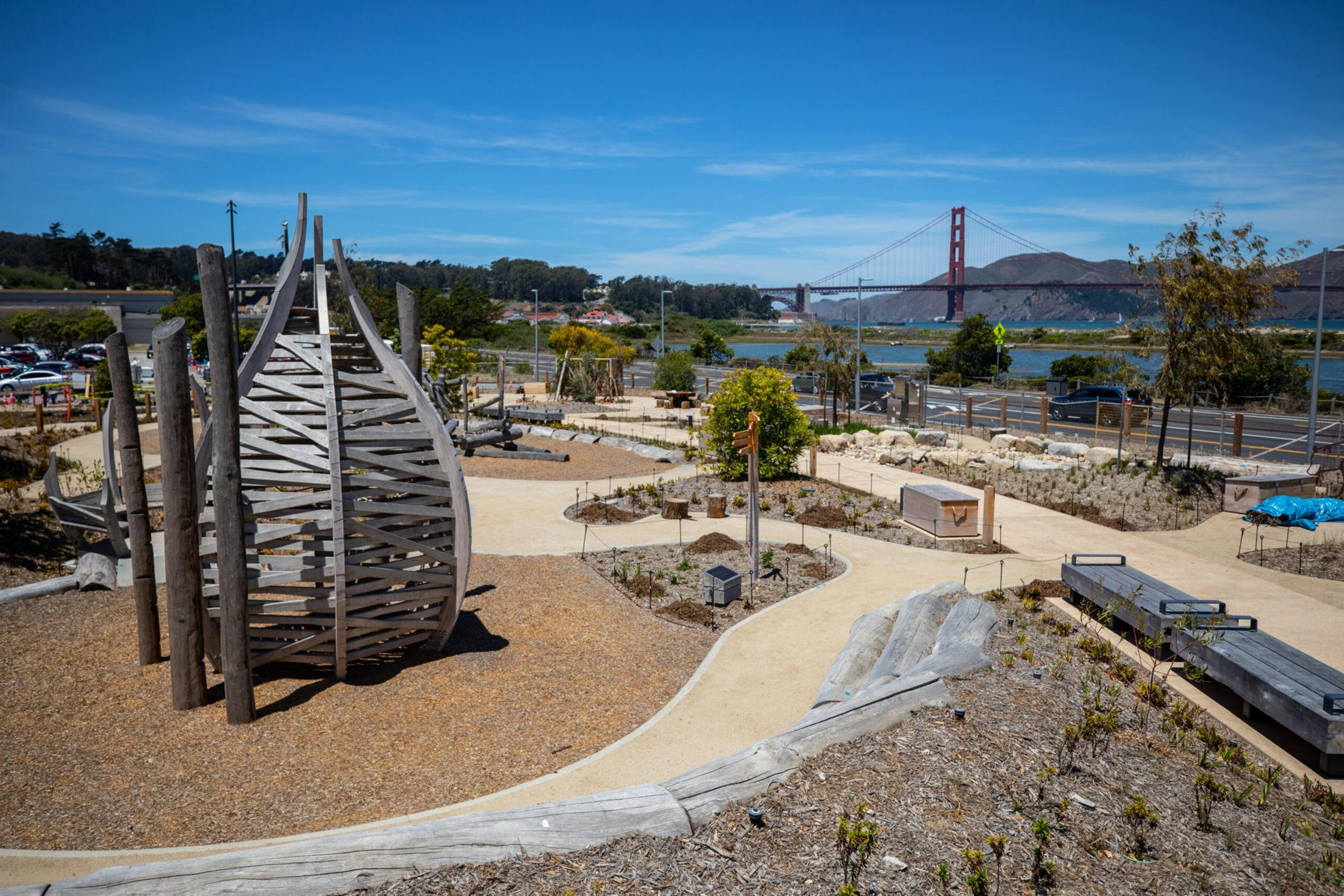 A landscaped park with winding paths, a wooden climbing structure, benches, and plants is set against a backdrop of water and the Golden Gate Bridge.