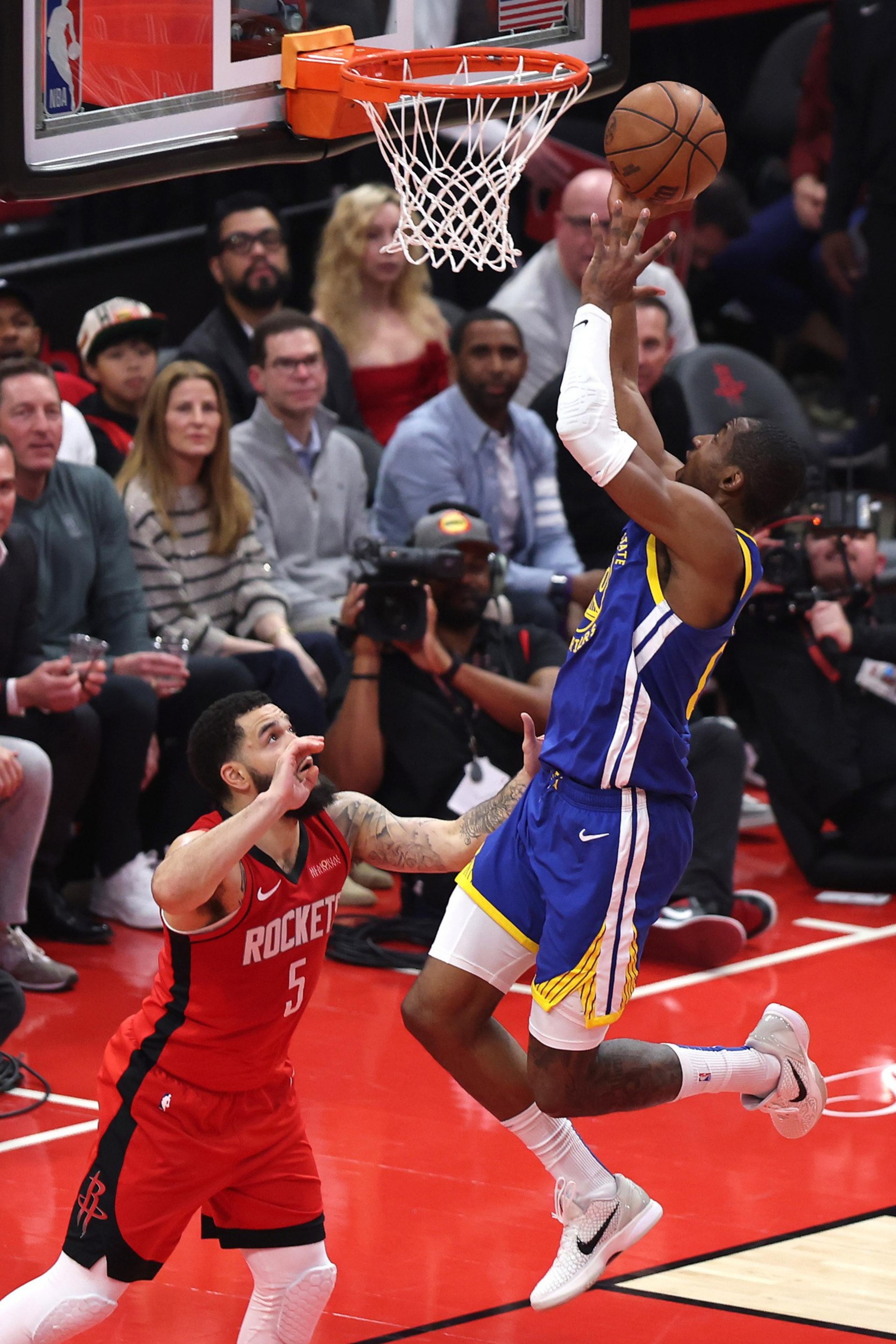 A basketball player in a blue jersey leaps towards the hoop for a shot, while a player in a red jersey attempts to defend. A crowd watches in the background.