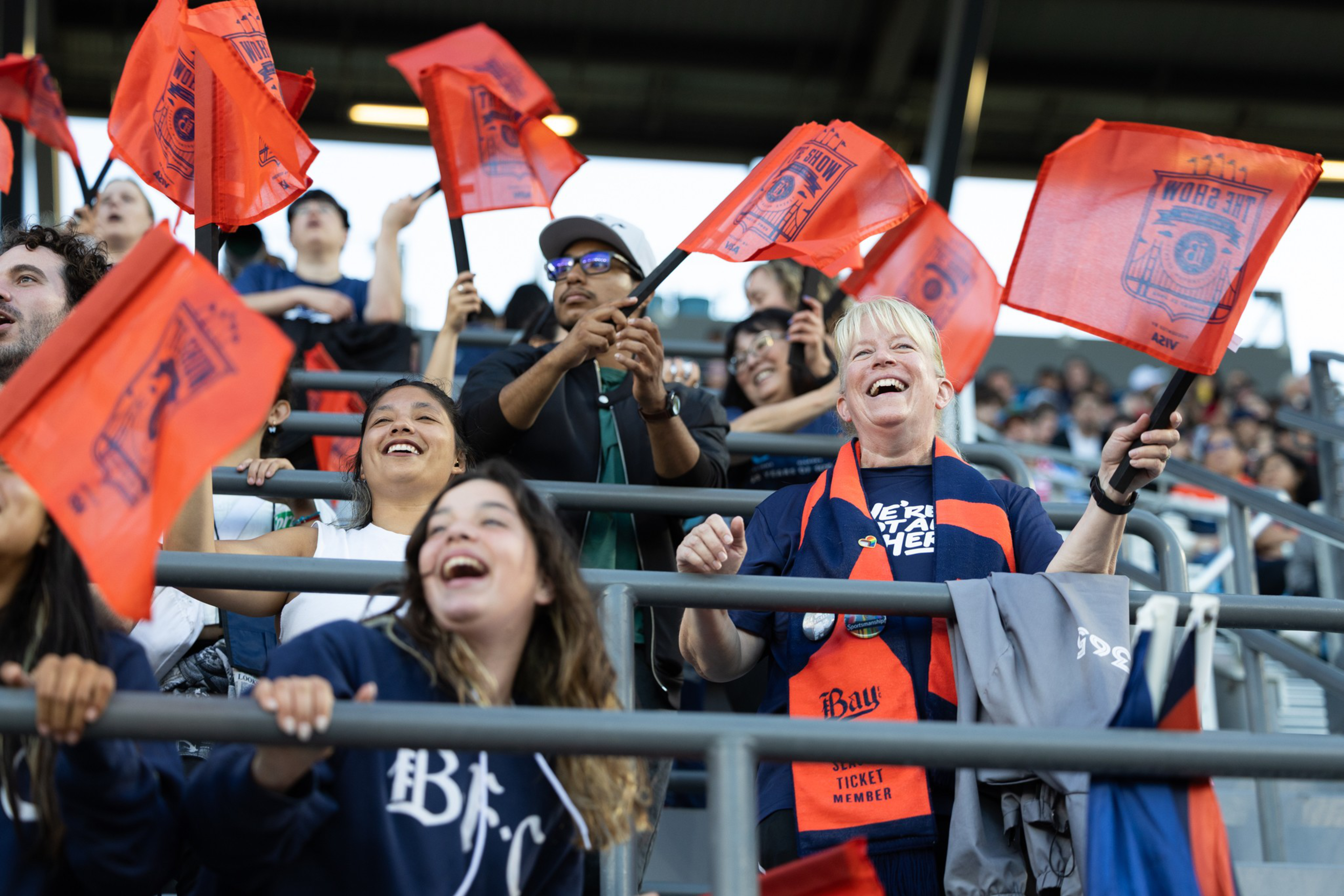 A crowd of fans wave orange flags and cheer excitedly in a stadium, showing support with scarves and smiles.