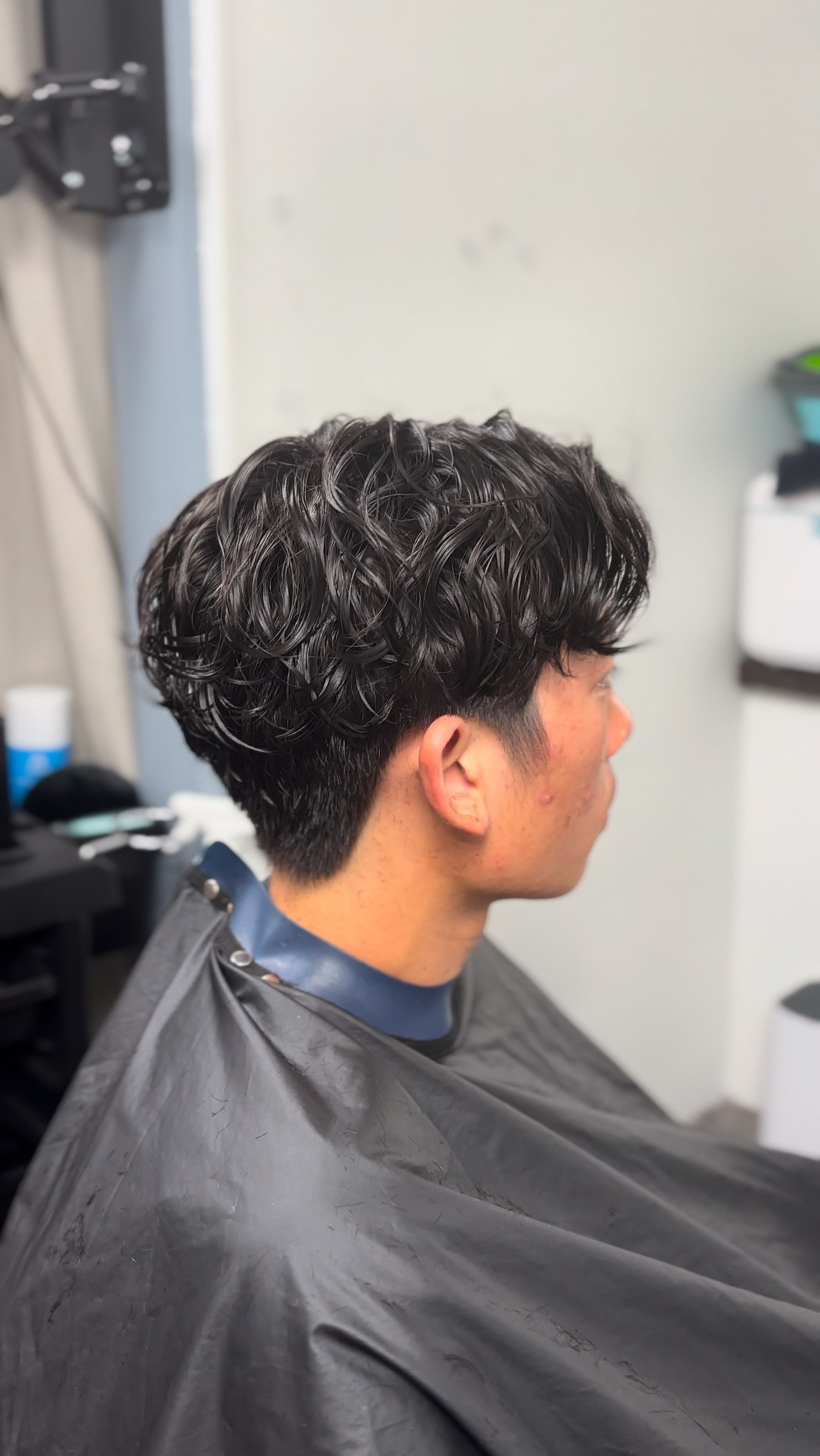 A person with dark, wavy hair is sitting in a salon chair, wearing a black haircut cape. The hair is neatly styled with a short trim on the sides.