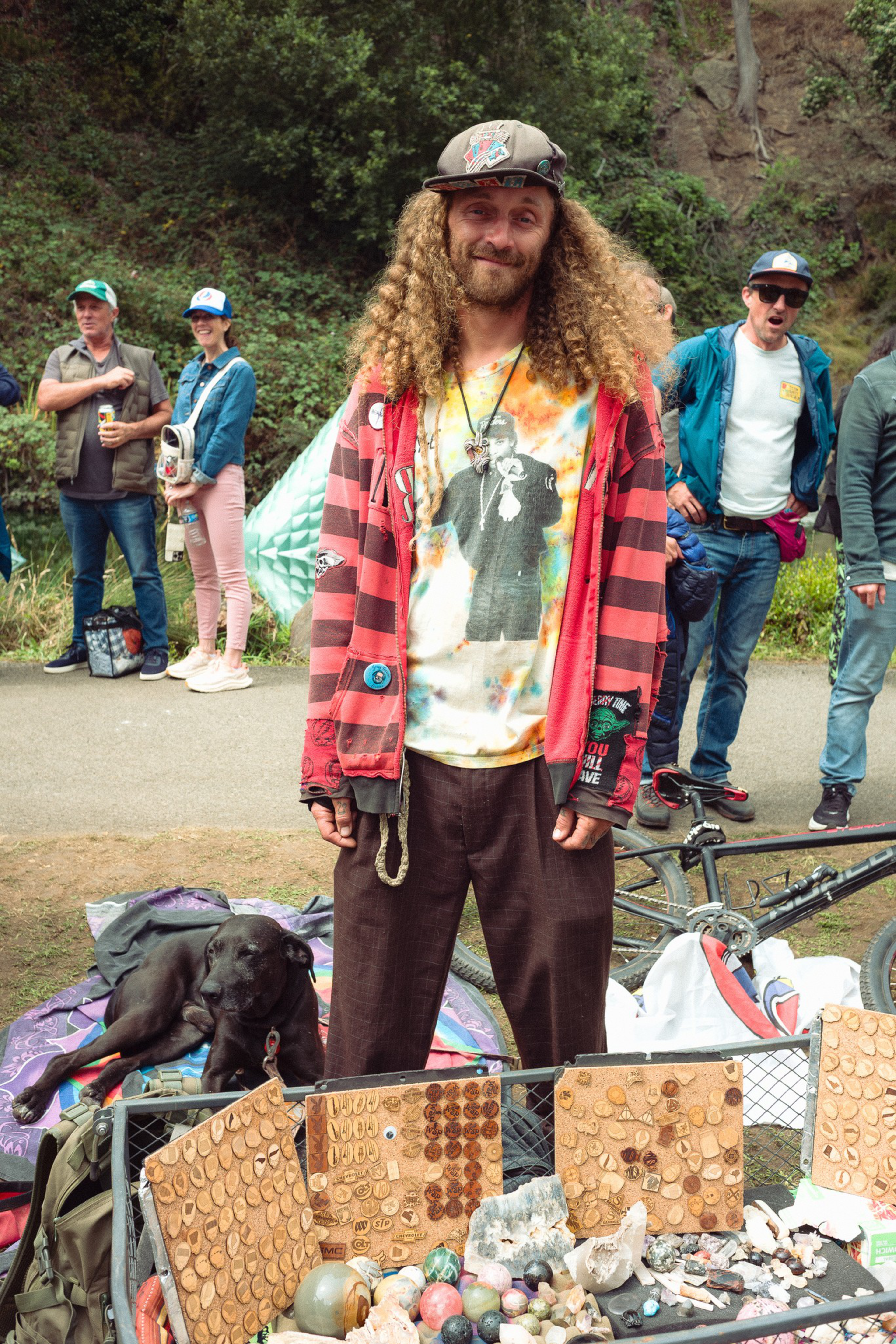 A man with long curly hair and a cap stands behind a display of rocks and coins, with a black dog lying on a blanket nearby and several people in the background.