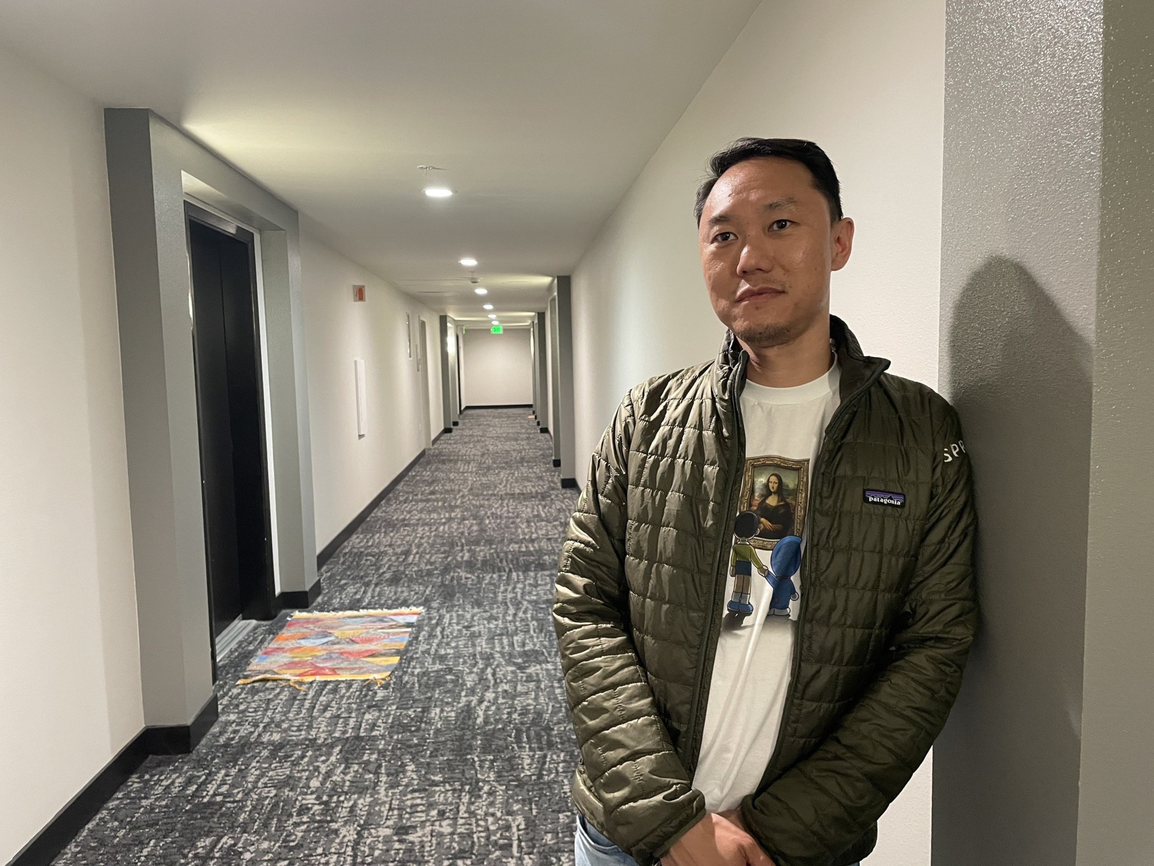 A man in a green jacket and a white T-shirt with a Mona Lisa print leans against a hallway wall with carpeted floors and closed doors.