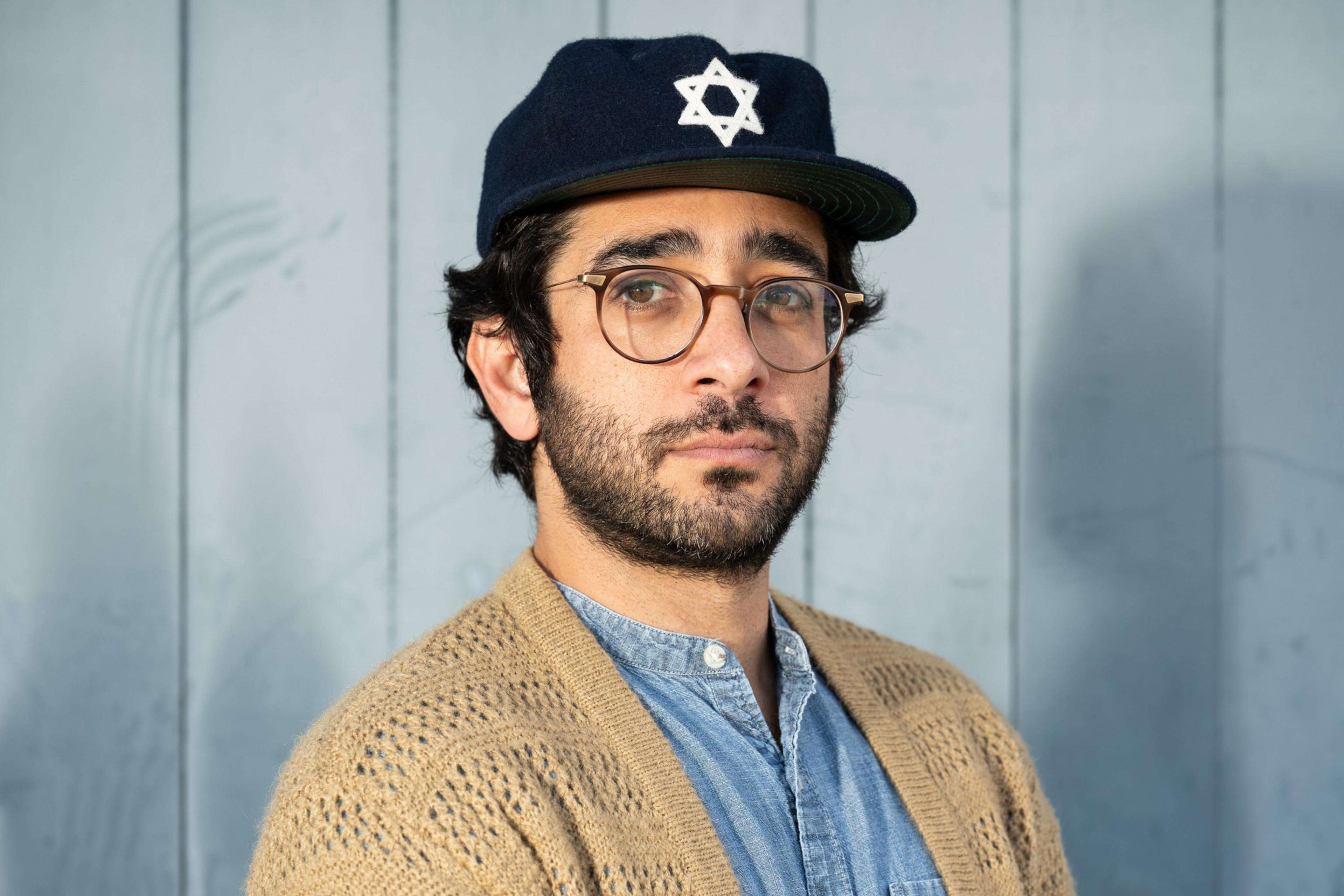 A person wearing glasses and a navy cap with a Star of David symbol is standing against a light blue wooden background, dressed in a denim shirt and a tan knit sweater.
