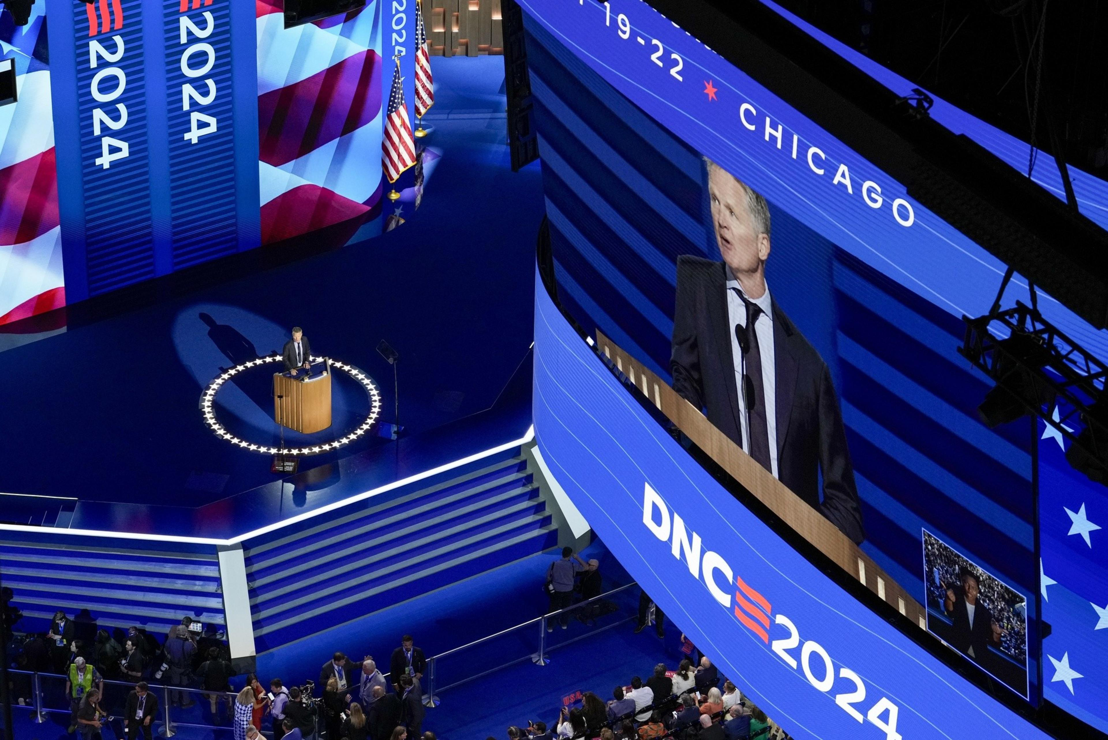 A speaker stands at a podium on a blue-lit stage, surrounded by stars. &quot;DNC 2024&quot; is displayed on screens, with an audience below and American flags visible.