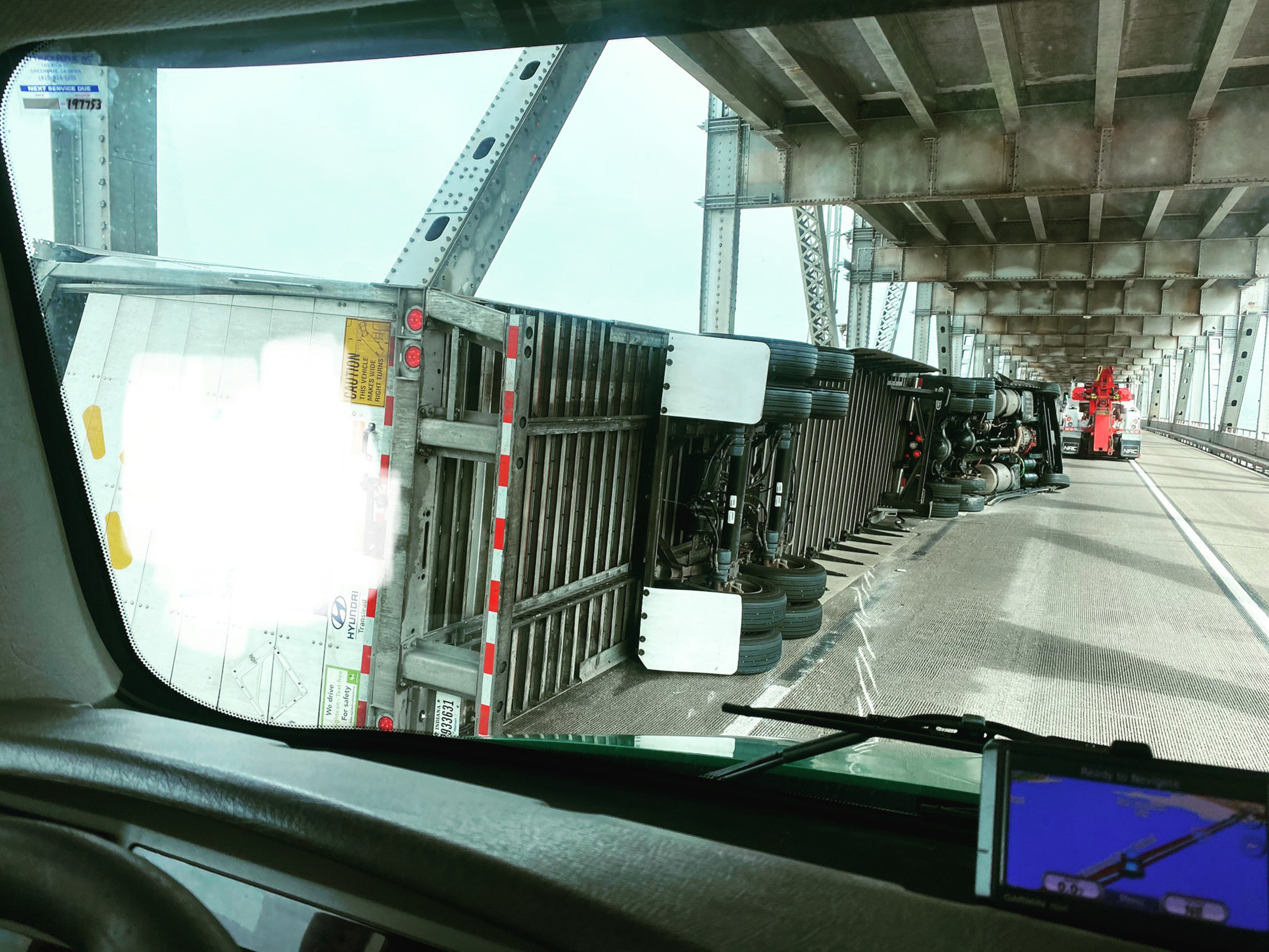 A large semi-truck trailer is overturned on its side on a bridge, with a tow vehicle approaching from behind to assist.