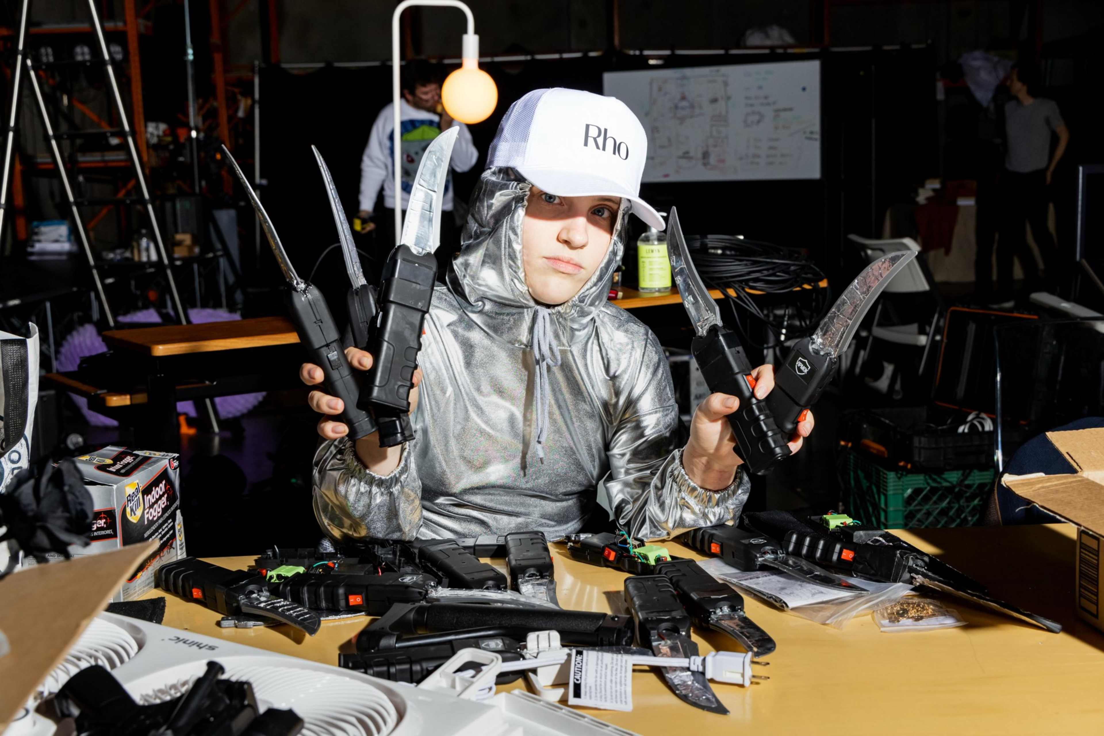 A person in a silver hoodie and white cap holds multiple large knives with black handles, sitting at a table cluttered with knives, gun replicas, and various items.