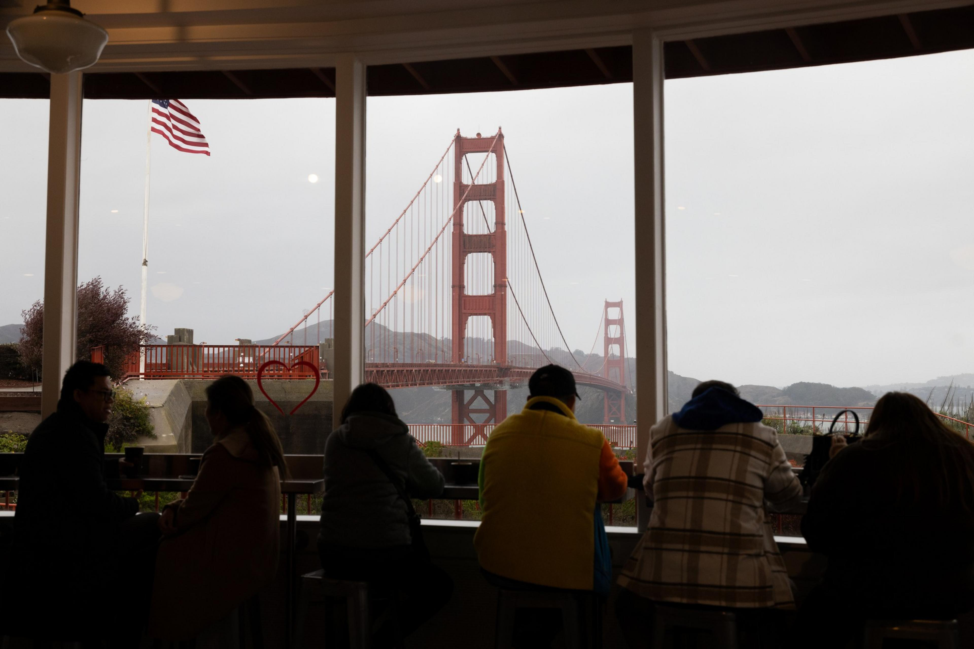 People sit inside a café, silhouetted against large windows framing the Golden Gate Bridge and an American flag outside, with overcast skies.