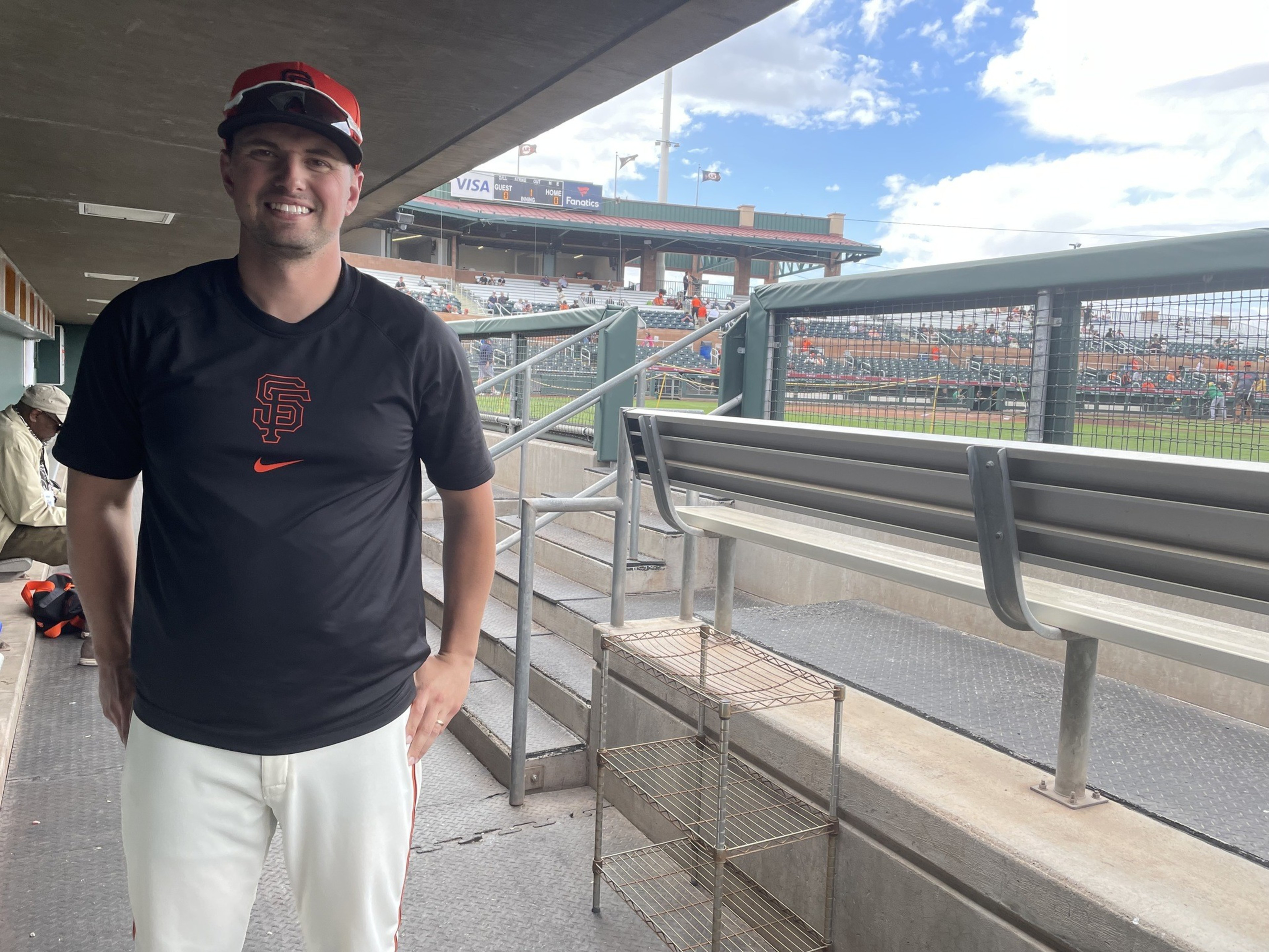 A person wearing a black San Francisco Giants shirt and cap stands in a baseball dugout, smiling. The field and stands with fans are visible behind them.