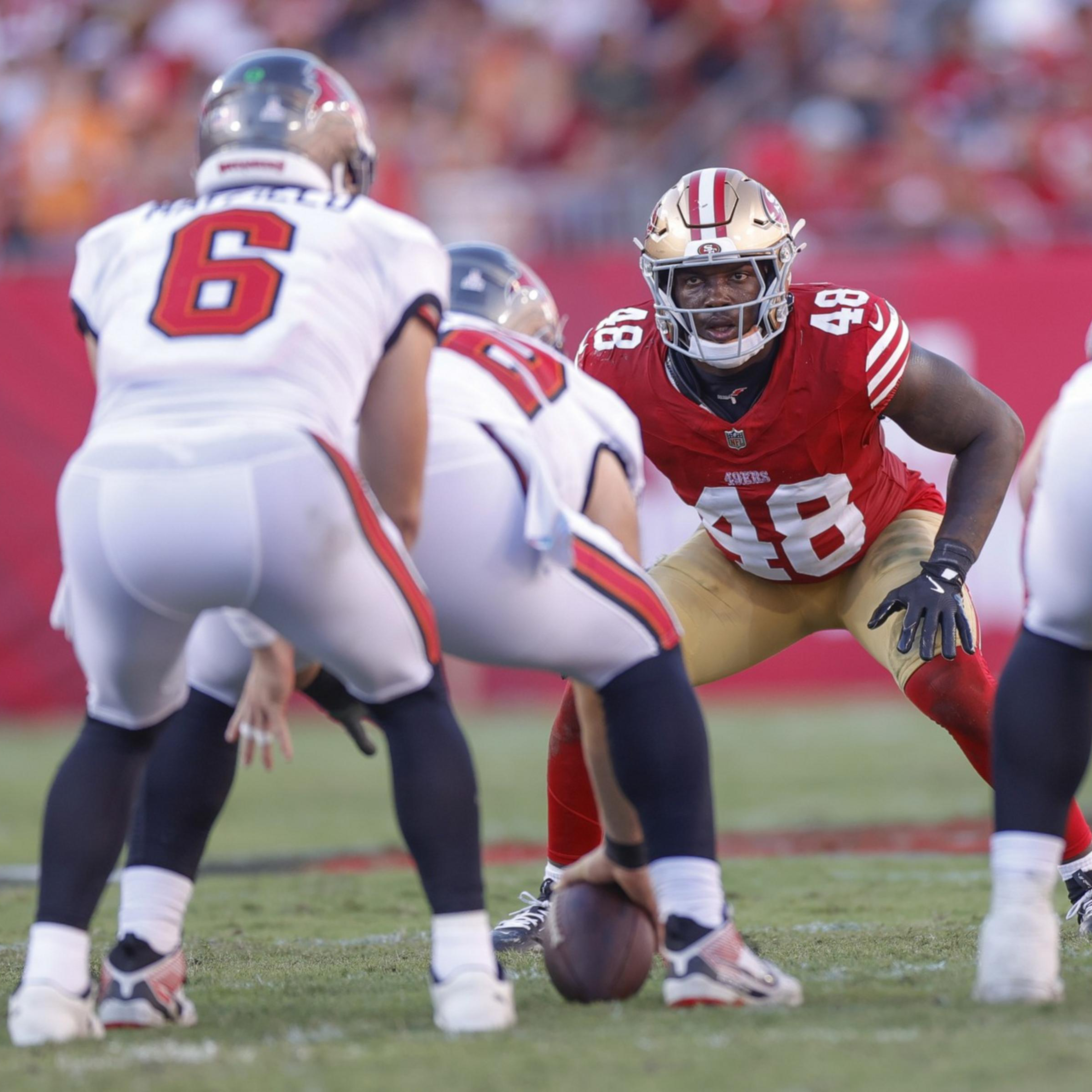 A football player in red and gold (#48) crouches in a defensive stance facing opponents in white uniforms, one holding the ball ready to snap.