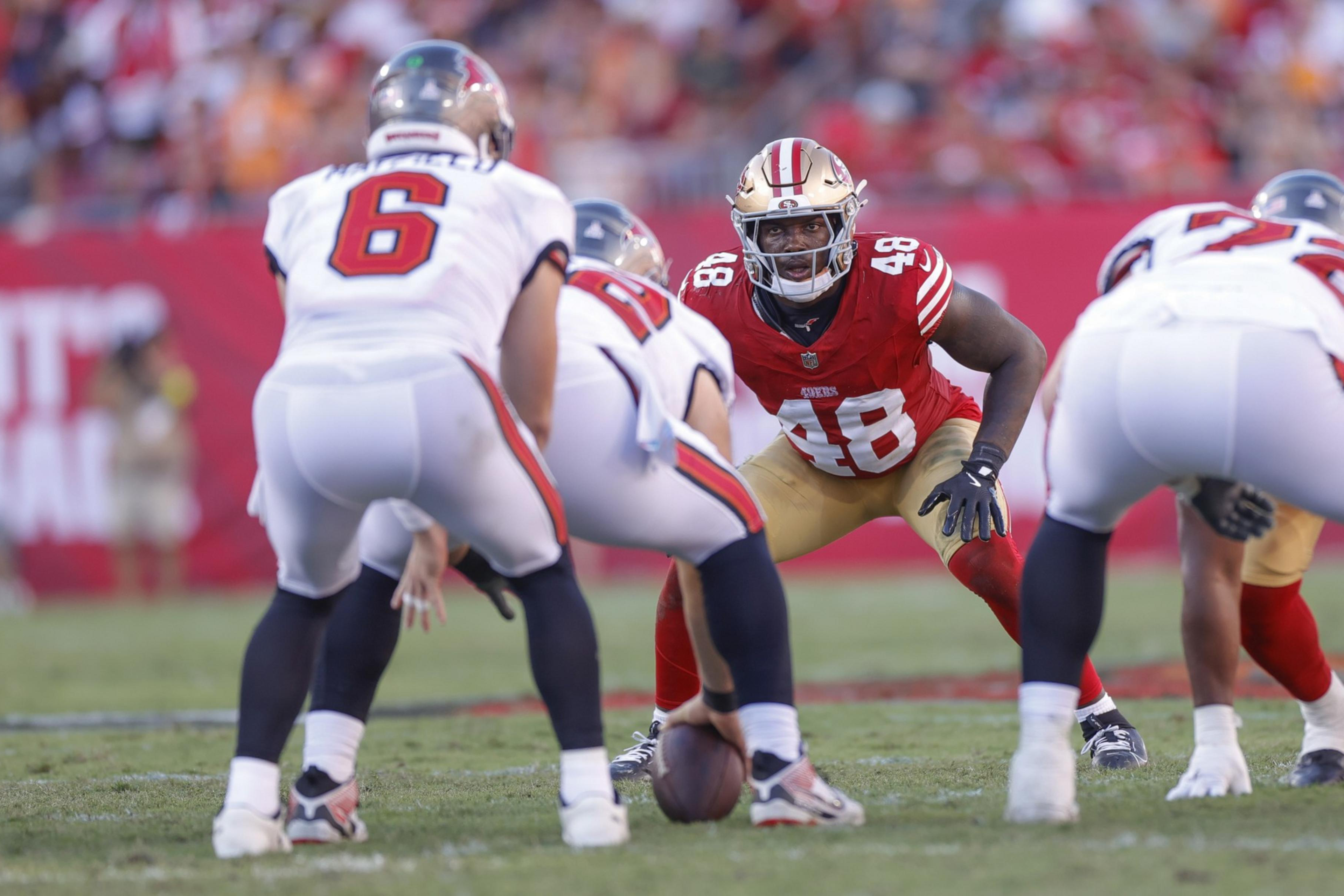 A football player in red and gold (#48) crouches in a defensive stance facing opponents in white uniforms, one holding the ball ready to snap.