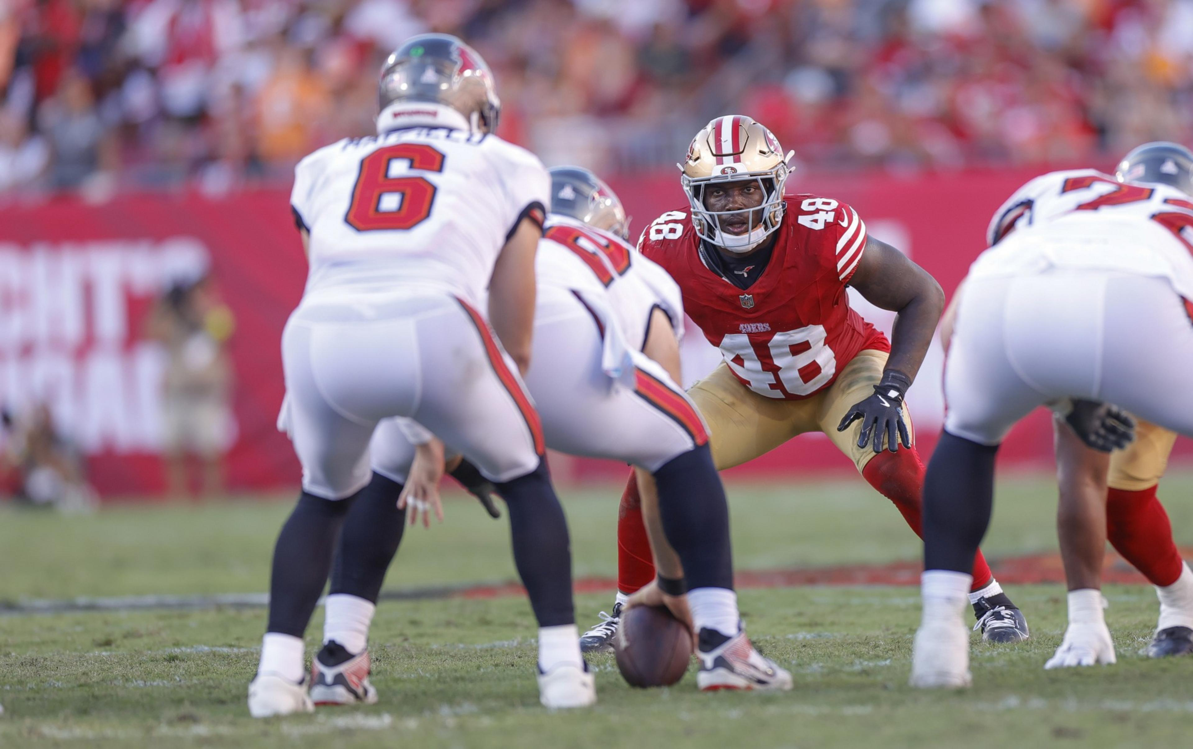 A football player in red and gold (#48) crouches in a defensive stance facing opponents in white uniforms, one holding the ball ready to snap.