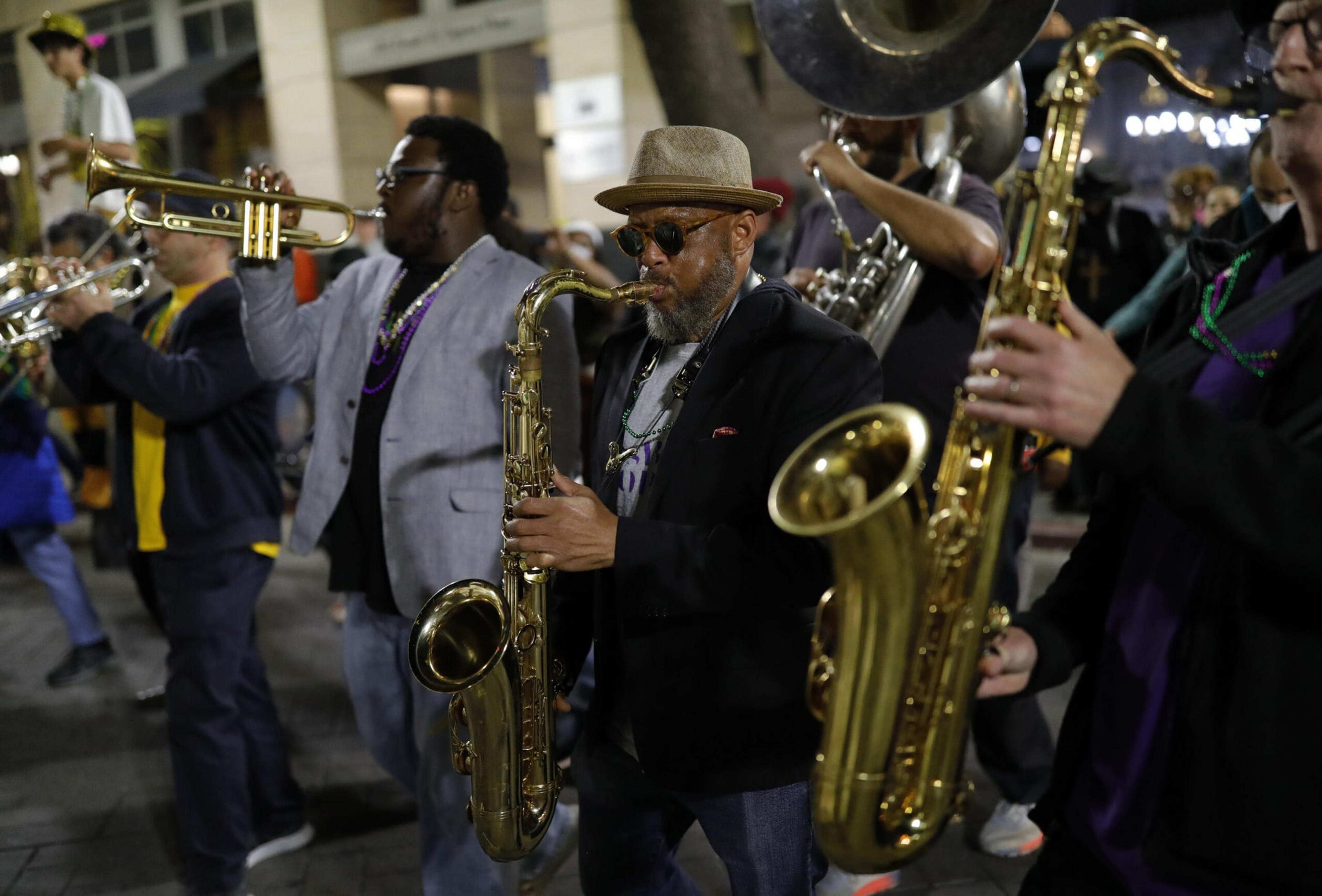 A lively brass band plays in a parade, with musicians wearing beads. They play trumpets, saxophones, and other instruments, dressed casually with hats and sunglasses.