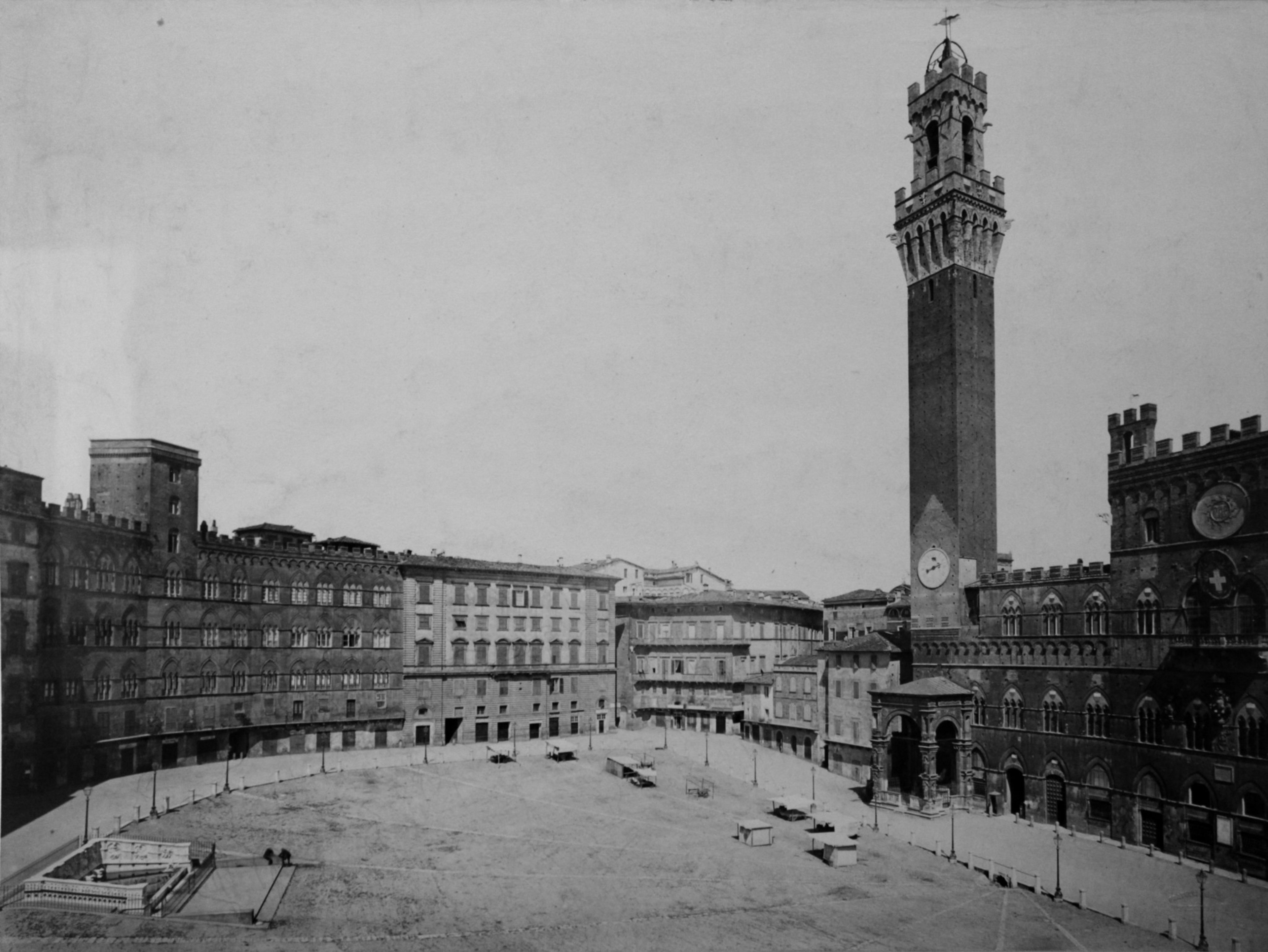 The image shows a large, empty medieval square with surrounding multi-story buildings and a tall clock tower with battlements on one side, typical of historic Italian architecture.