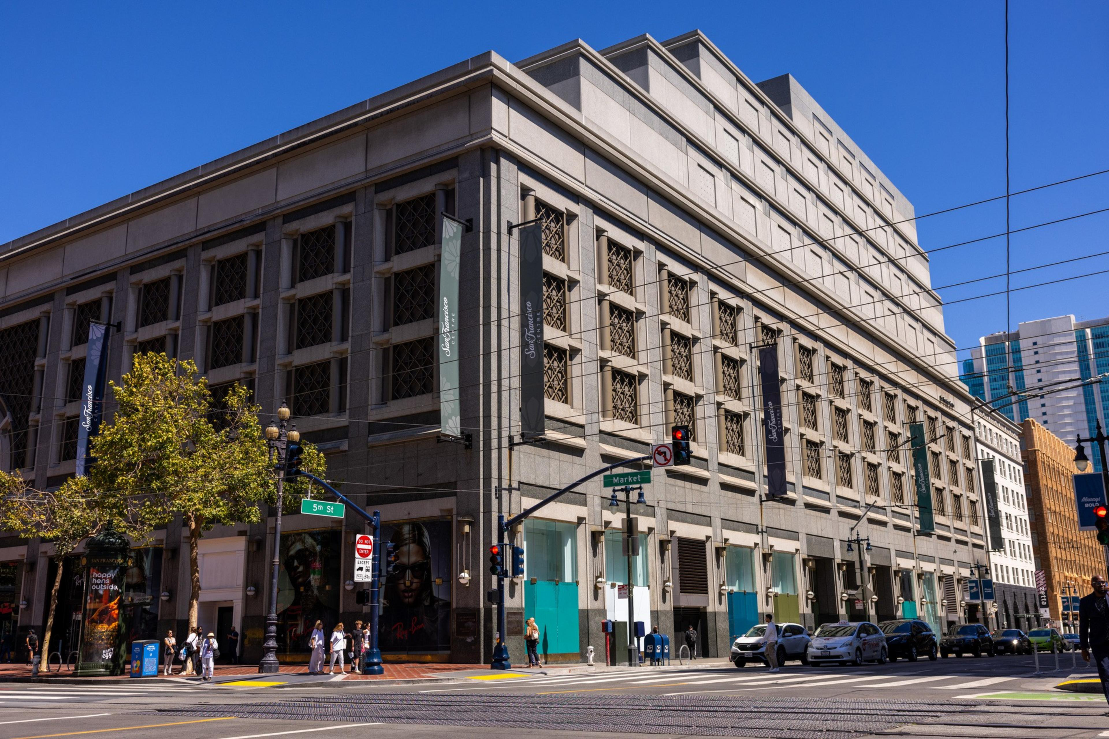 A large, multi-story concrete building stands at the corner of 5th St and Market St with cars parked along the street and pedestrians on the sidewalk.