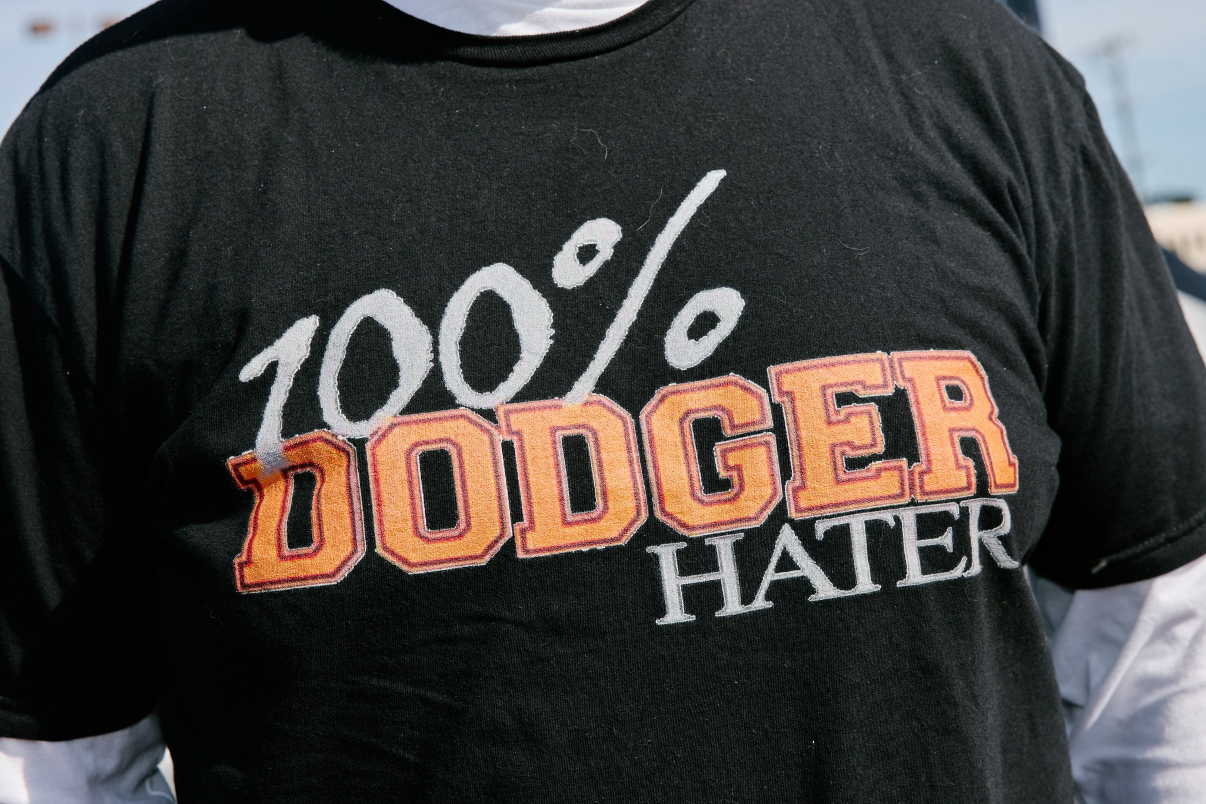 A black T-shirt with bold orange and white text reads “100% DODGER HATER,” worn over a long white-sleeve shirt.