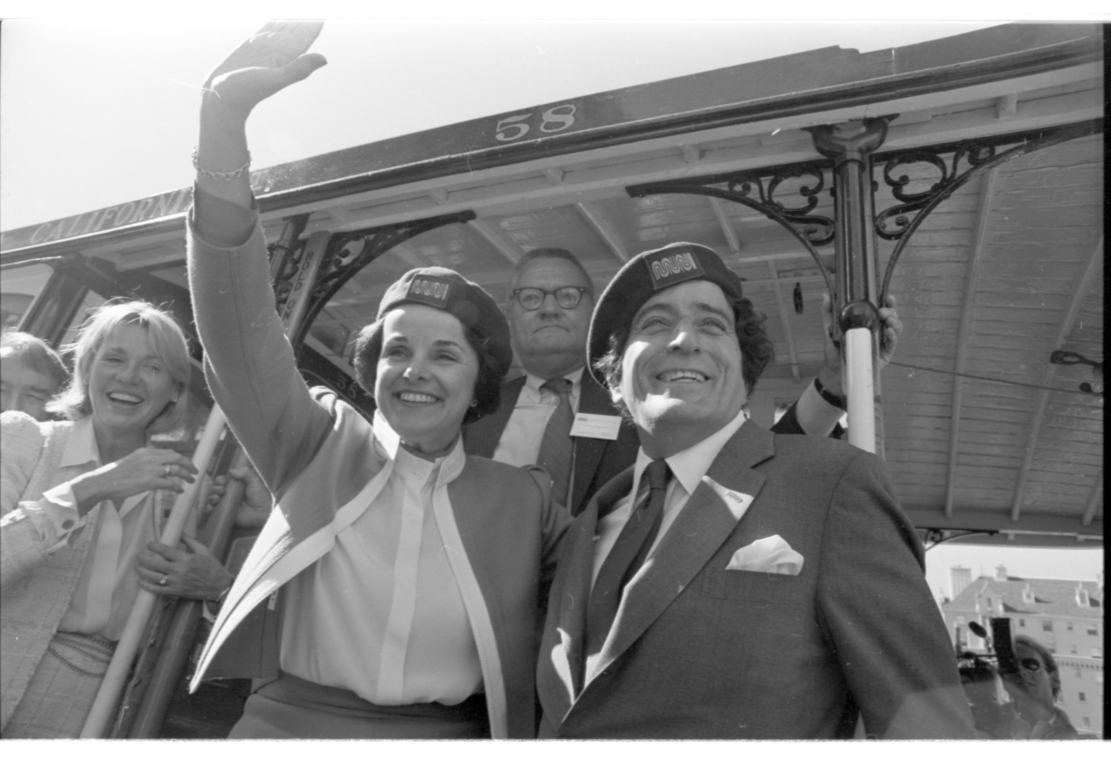 A smiling man and woman wearing matching hats wave from a cable car, with two additional people visible behind them.