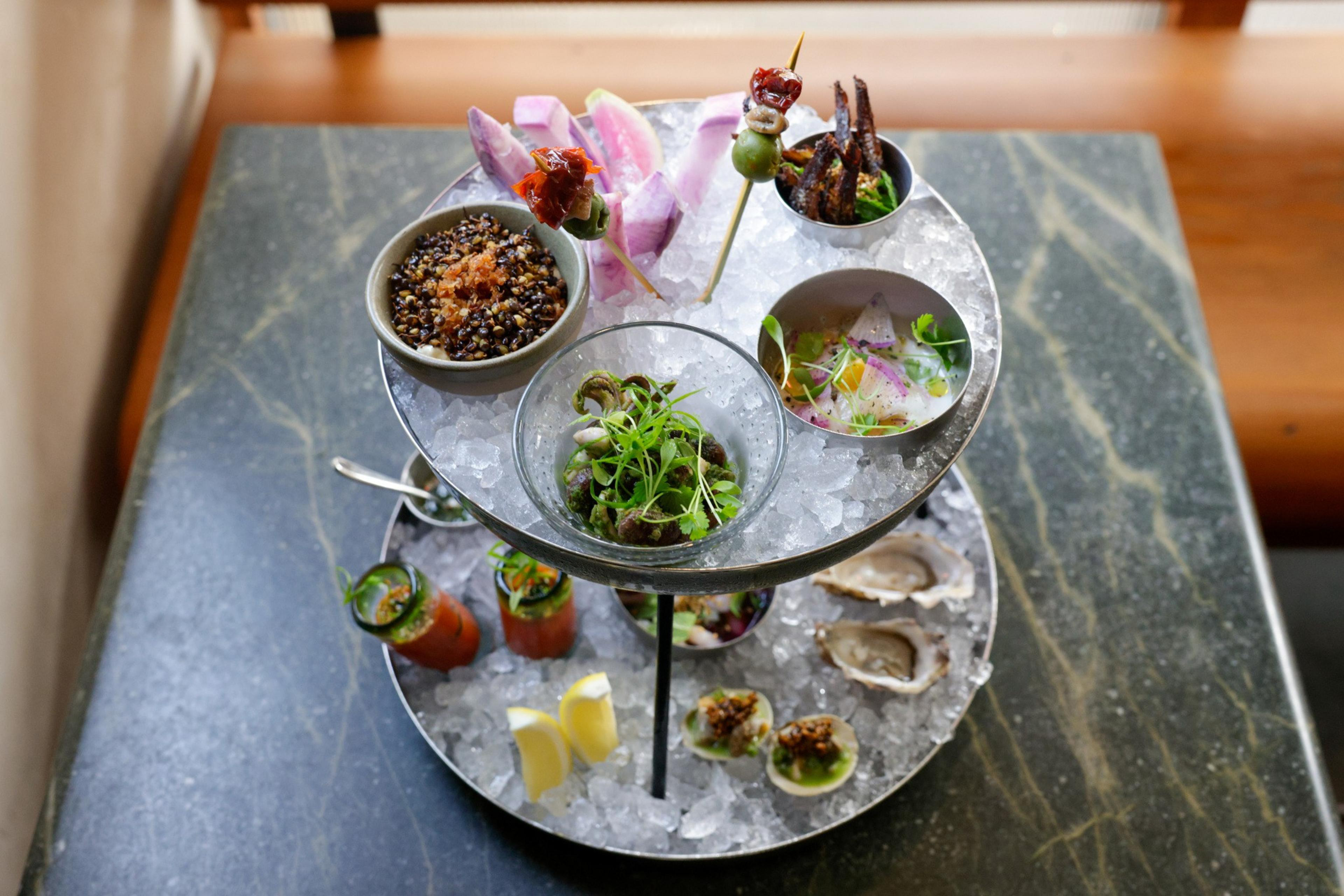A two-tiered serving tray holds an array of gourmet appetizers on a marble table.