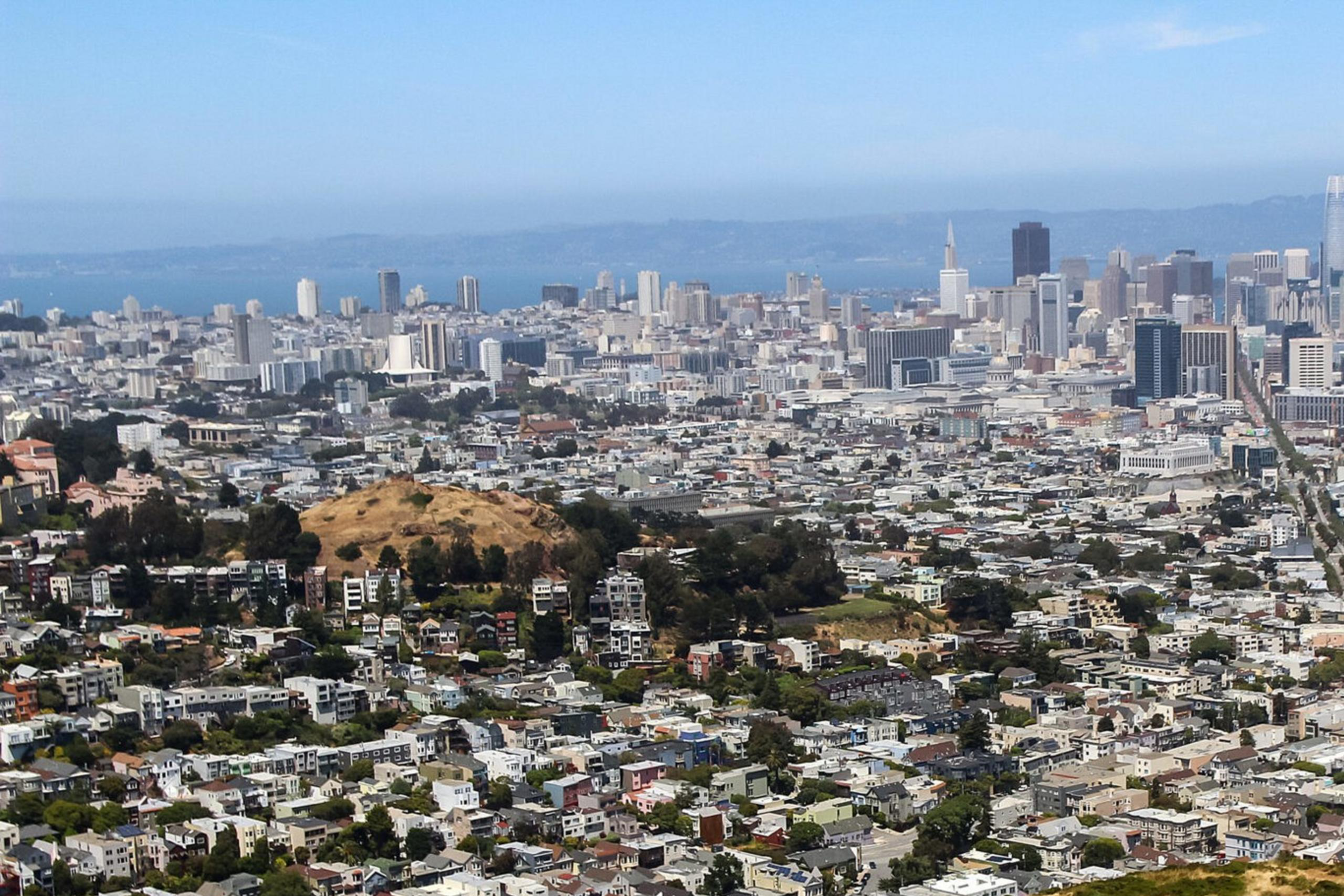 A cityscape with tall buildings in the distance, viewed from above a windy road on a hill, showing a mix of residential and commercial areas under a clear sky.
