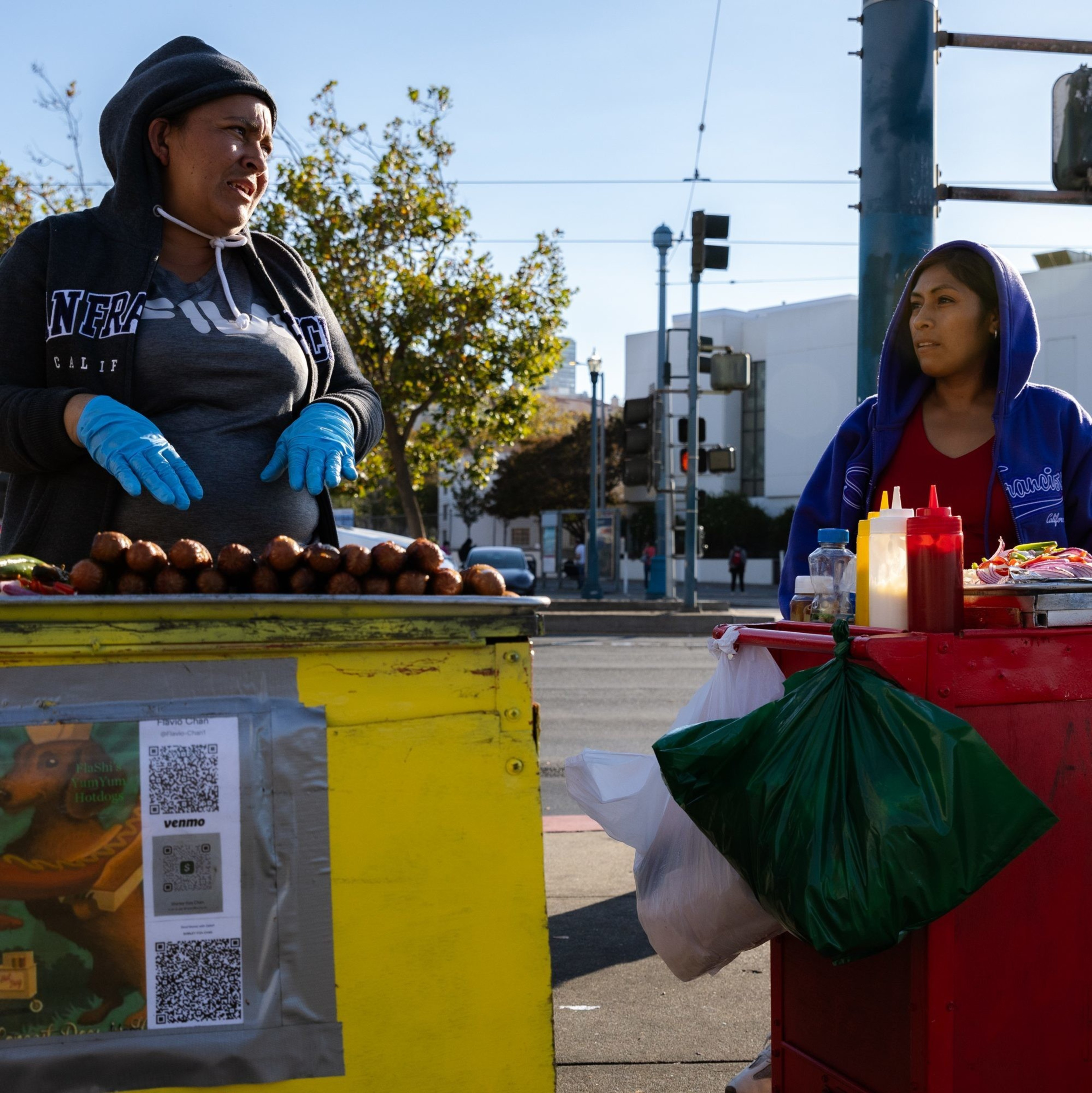 Two women stand behind street food carts, one in a gray hoodie and blue gloves, the other in a blue hoodie. They're selling food with condiments and QR codes visible.