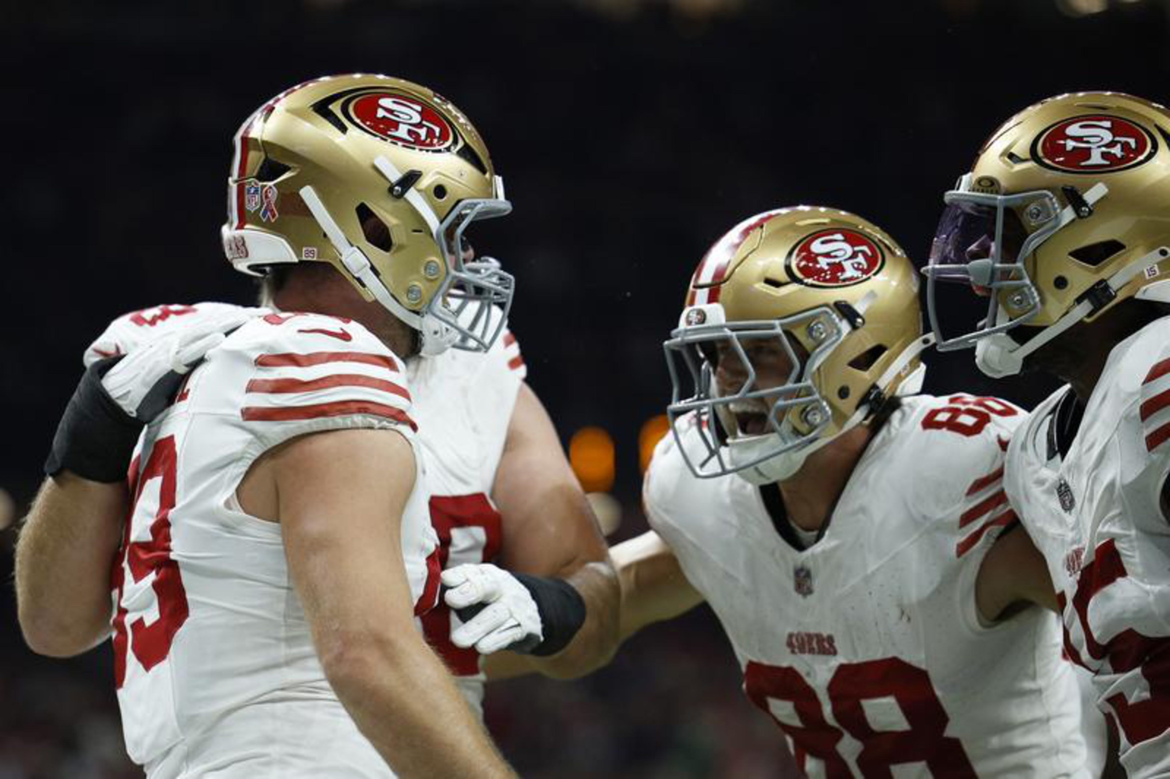 Three San Francisco 49ers players in white jerseys and gold helmets celebrate closely during a football game.