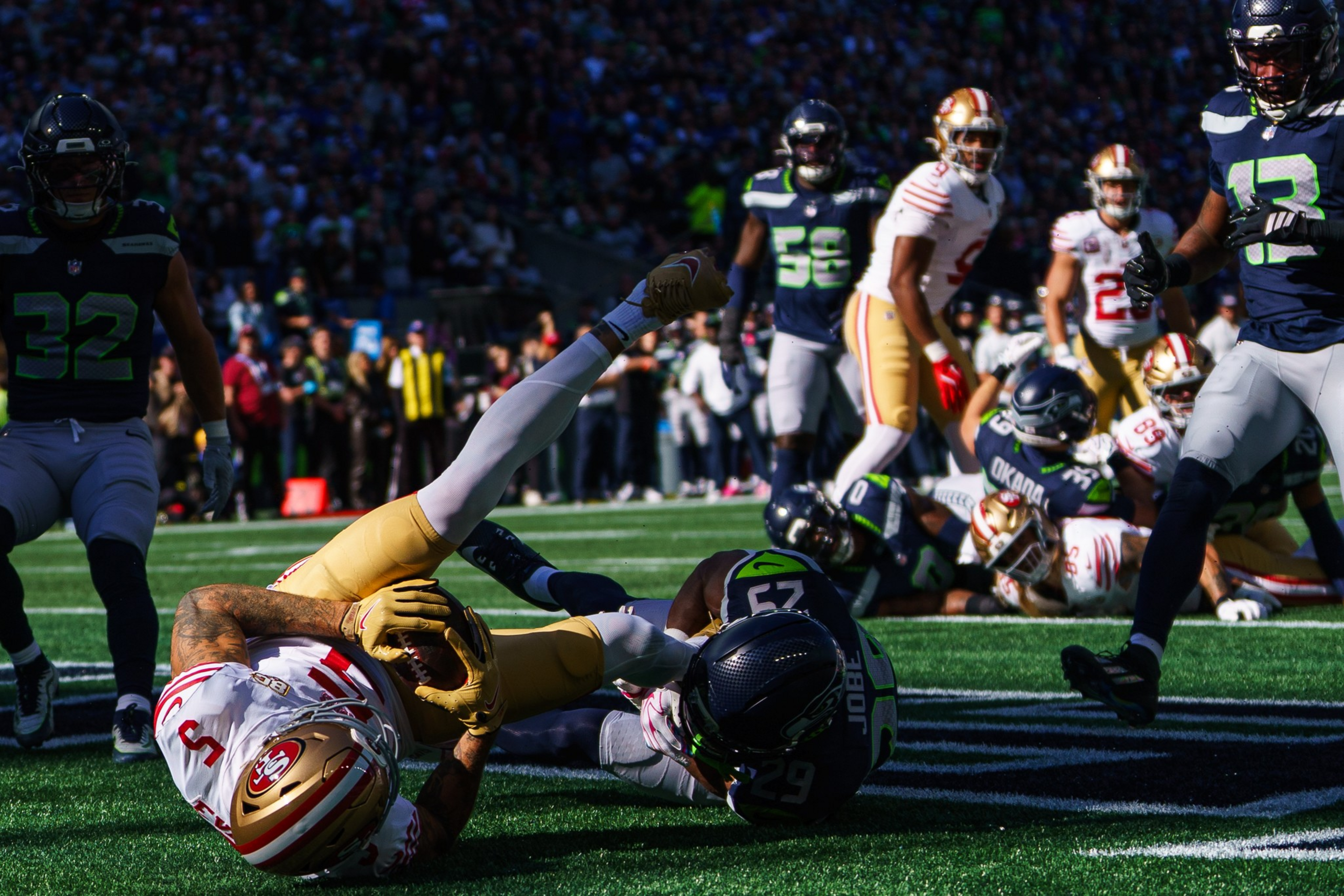 A San Francisco 49ers player is tackled by a Seattle Seahawks defender near the end zone, with other players and spectators visible in the background.