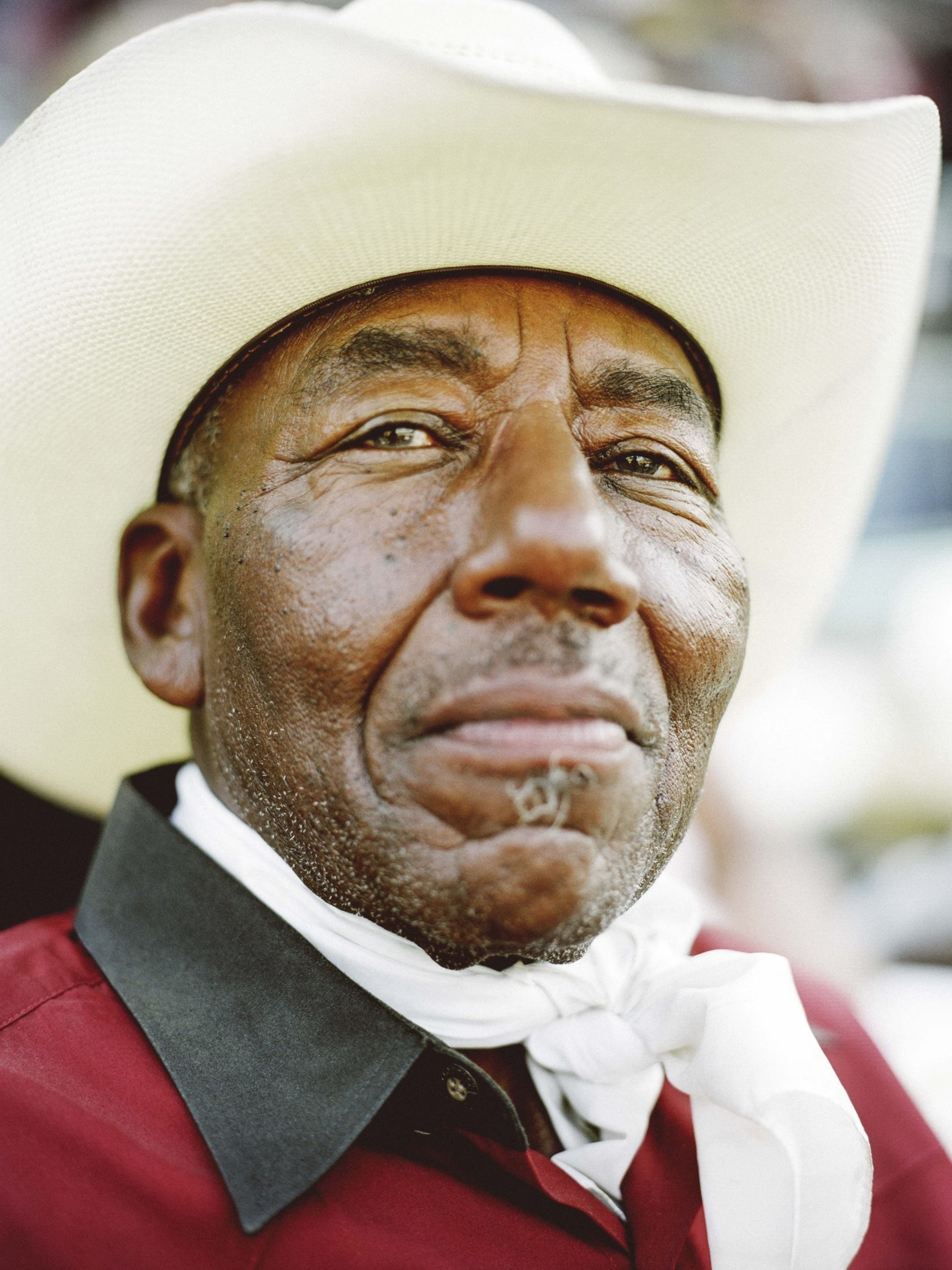 An elderly man wears a white cowboy hat, a red shirt with a black collar, and a white neckerchief, looking contently into the distance.