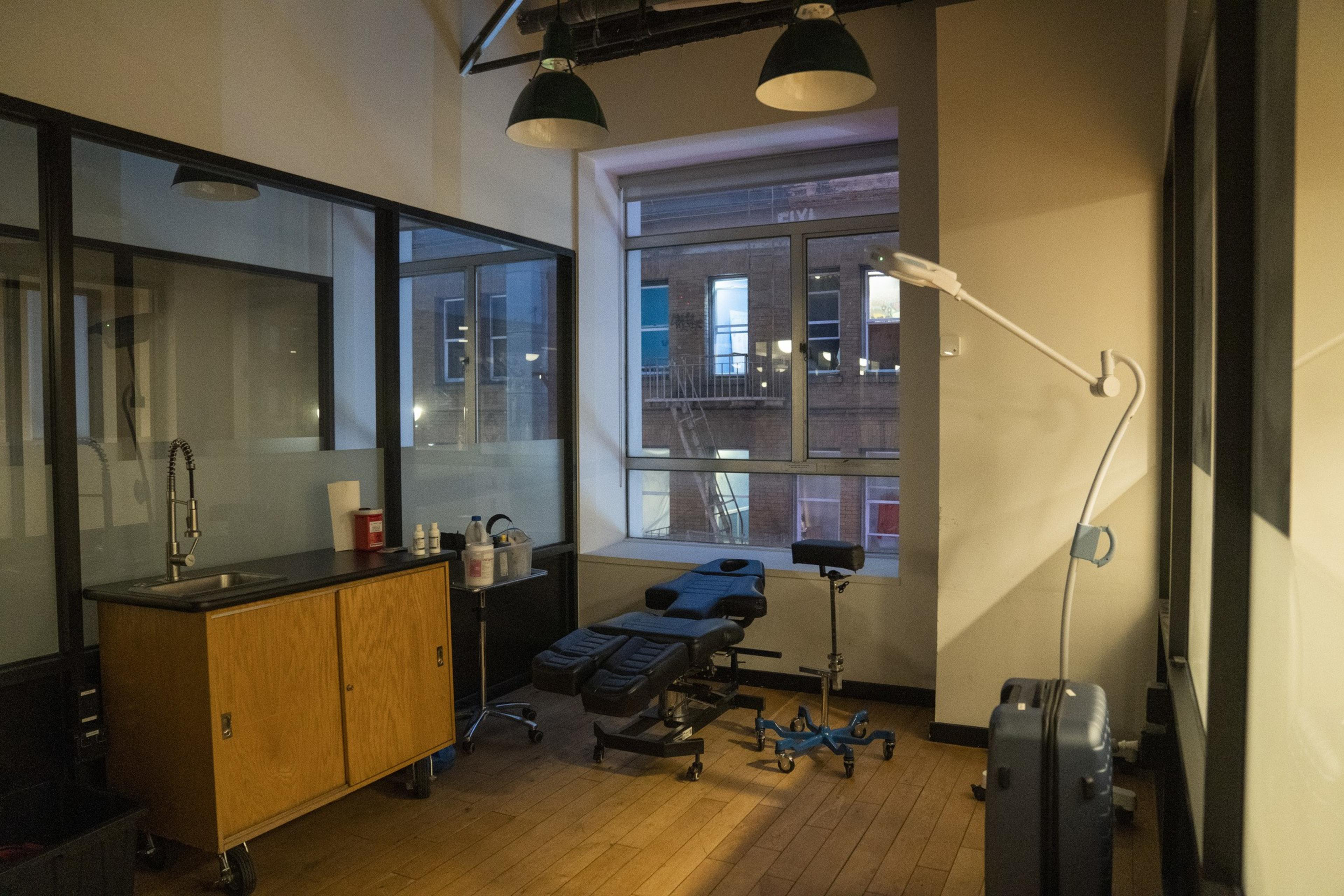 The image shows a room with a black adjustable chair, wooden cabinets, a sink, and a rolling cart with supplies. Large windows reveal a building outside.