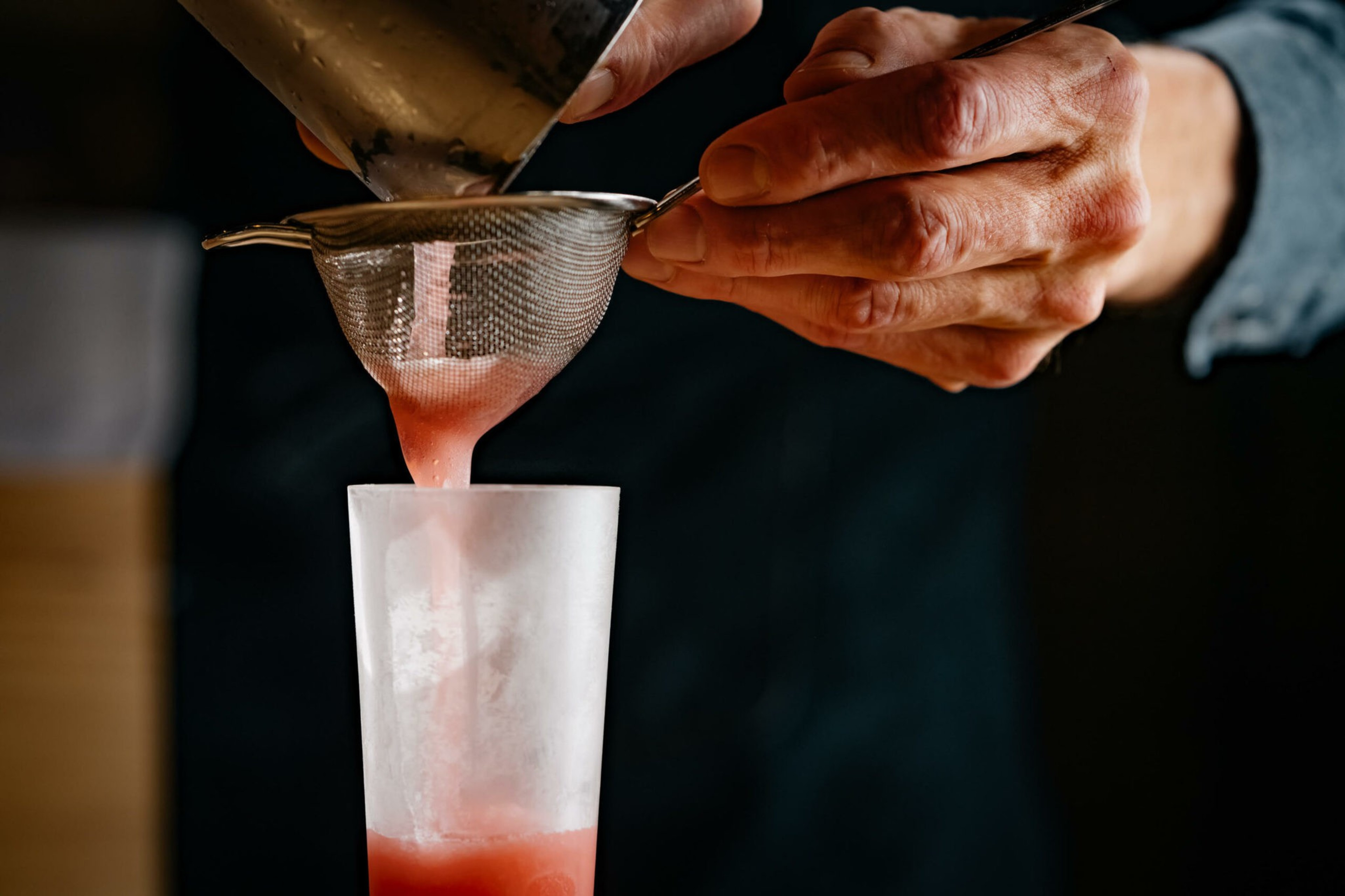 A person's hands are straining a rose-colored cocktail.
