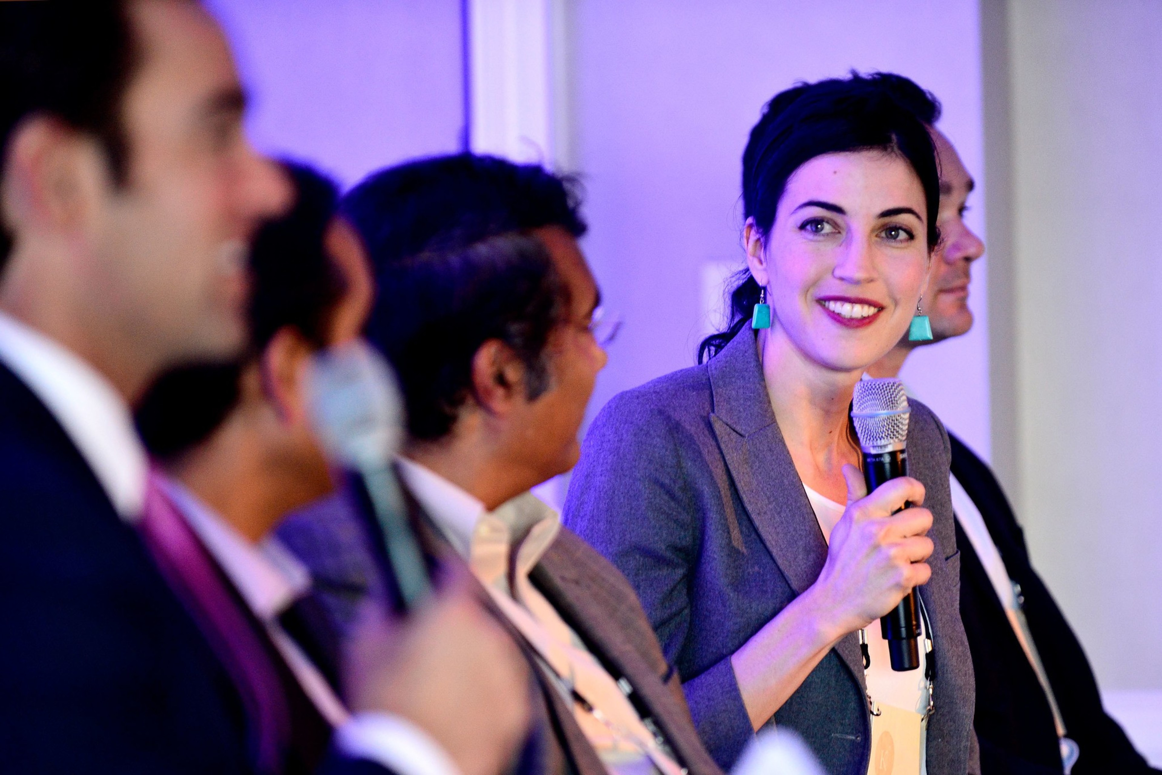 Three men can be seen in the foreground as OpenAI board member Tasha McCauley talks, holding a microphone, at a panel. To her right is another man on a panel.