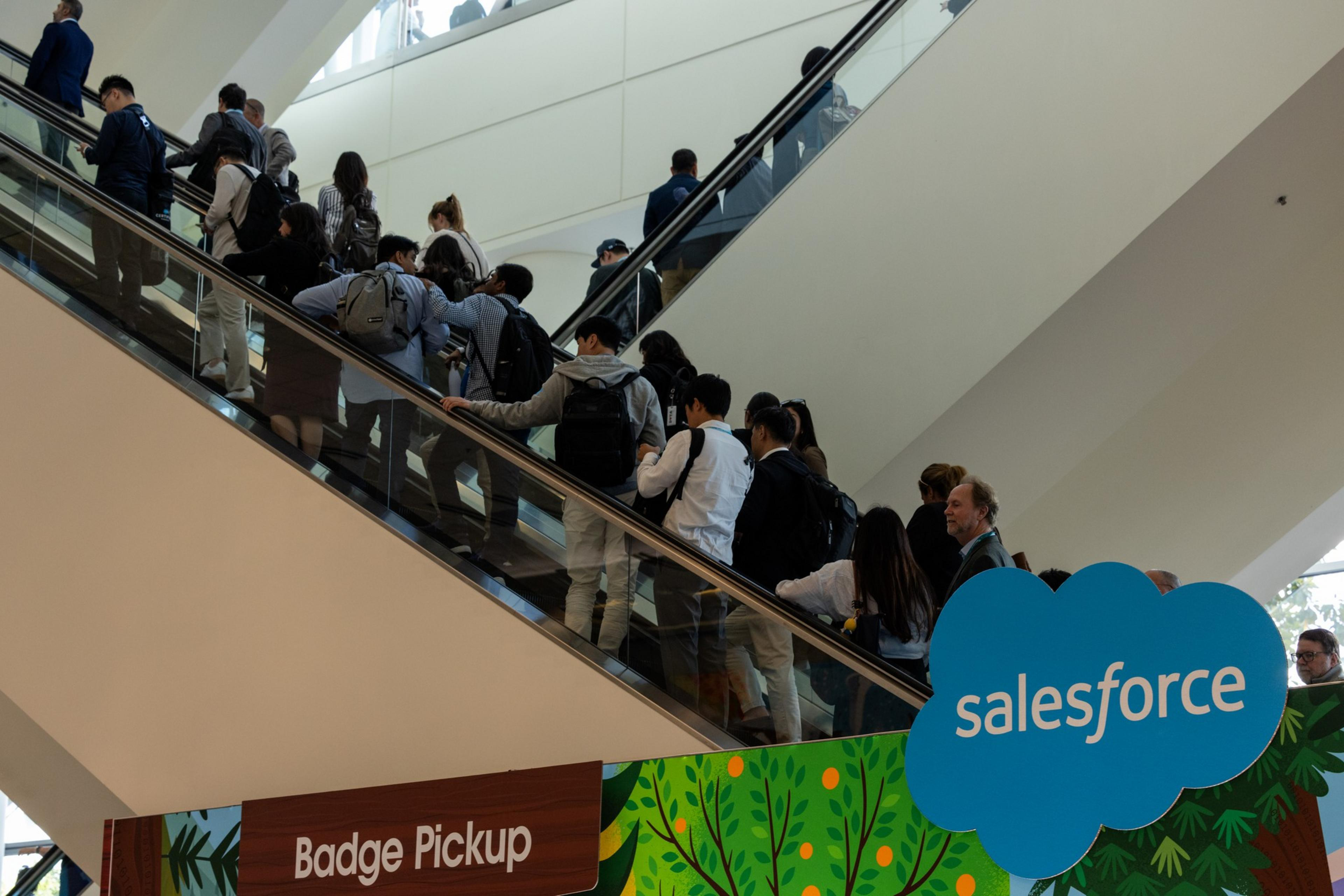 People with backpacks stand on an escalator in a line heading upward inside a building, near signs for "Badge Pickup" and "Salesforce."