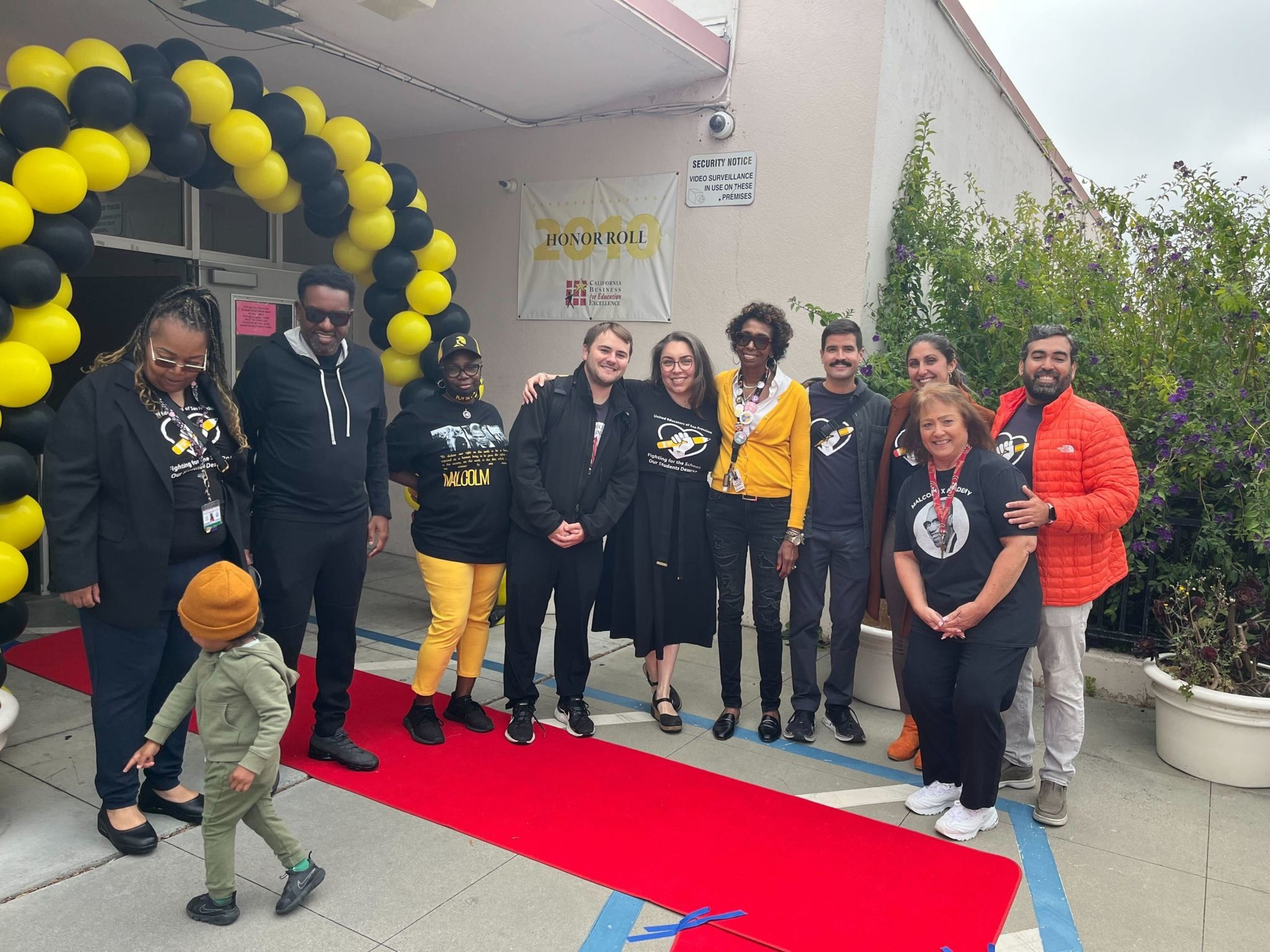 A group of eleven people are smiling and posing on a red carpet under a black and yellow balloon arch, with an "Honor Roll" sign and greenery in the background.