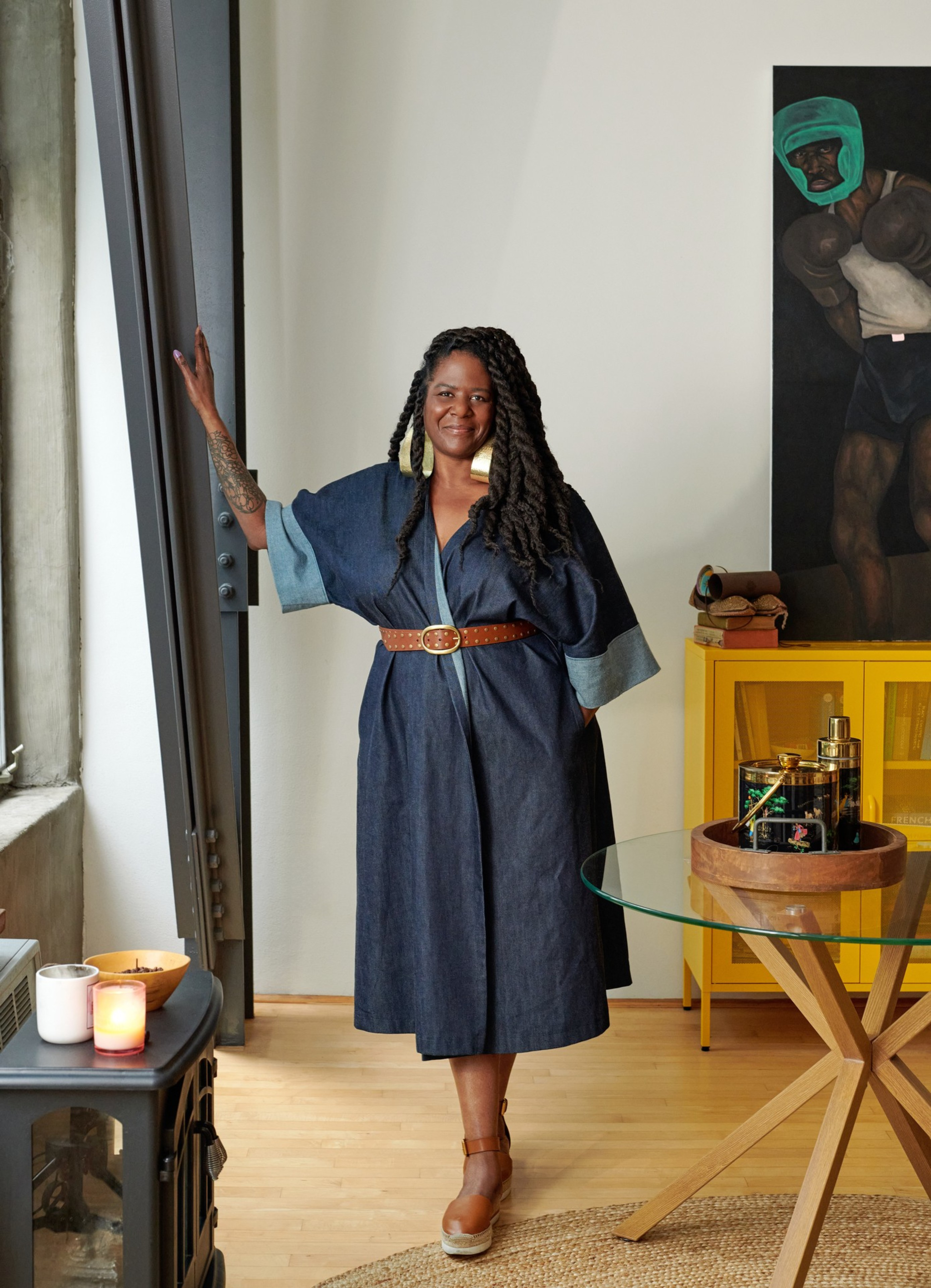 A woman in a denim dress stands by a window, smiling. Her hair is styled in long braids, and she is wearing large earrings. A painting and candles are visible nearby.