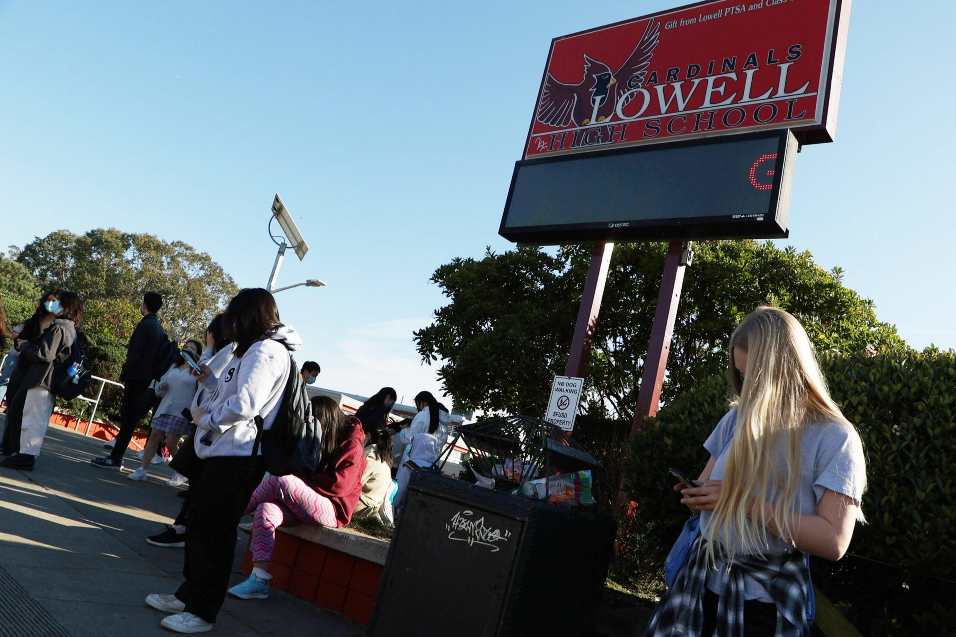 Students stand and sit near a sign that reads "Lowell High School Cardinals" on a sunny day, some looking at their phones or wearing masks.